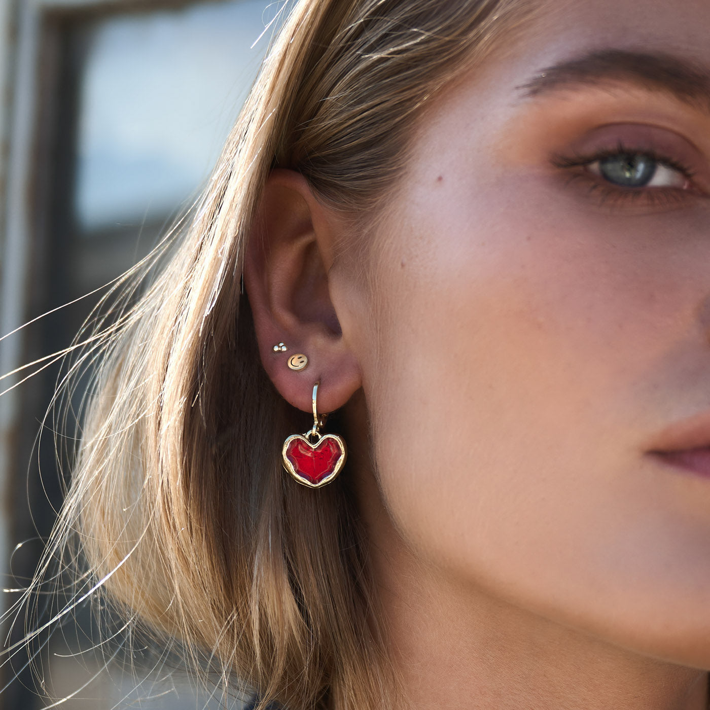 Dixie - Smiley Face Stud Earrings Stainless Steel - BECCASIN STORE - Gold-tone smiley face stud örhänge paired with hanging red heart smycke on light hair.