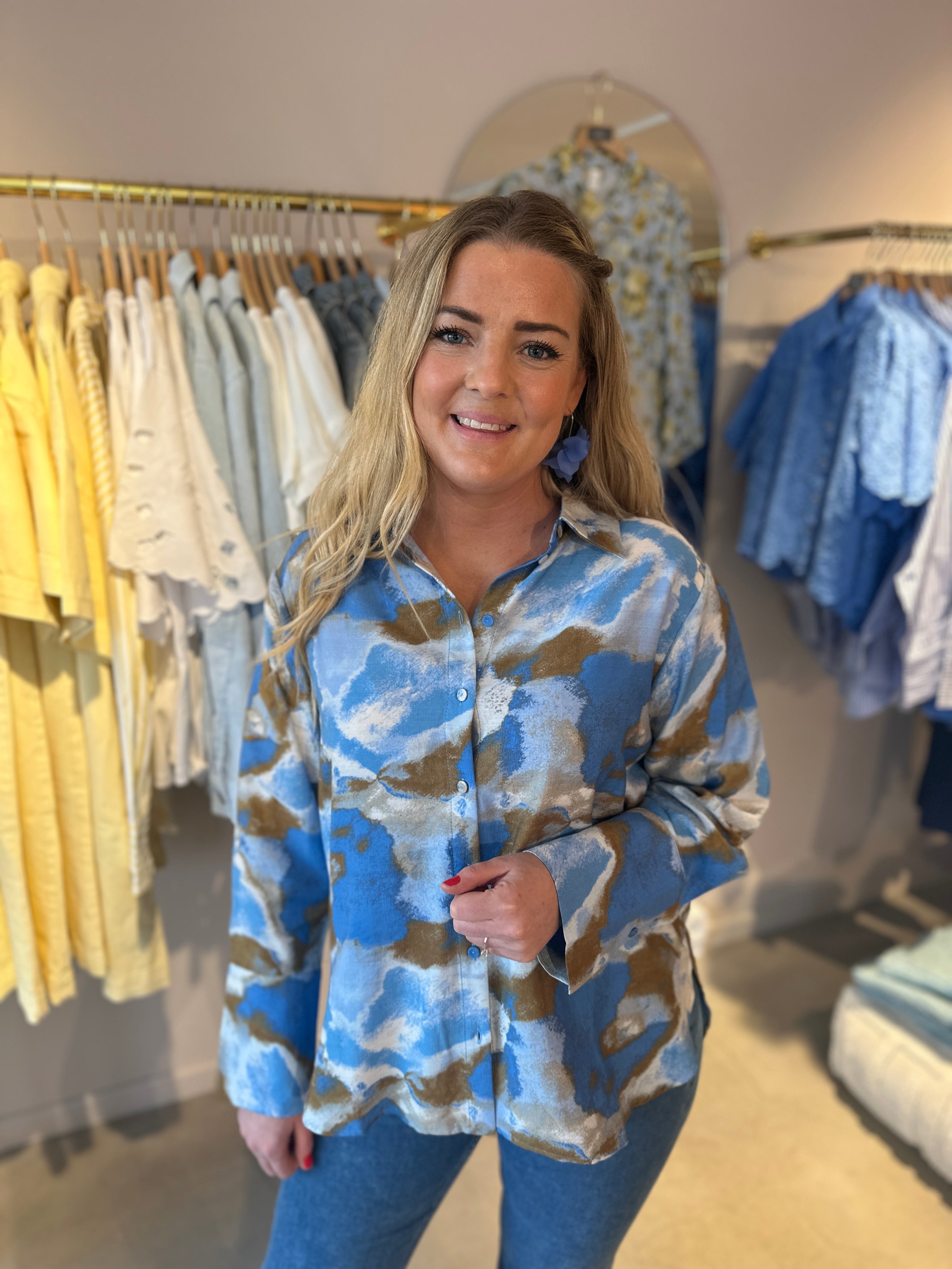 Kvinna i oversized skjorta med abstrakt mönster i blå, khaki och vitt, helbild framifrån i butiksmiljö med klädstativ i bakgrunden.
