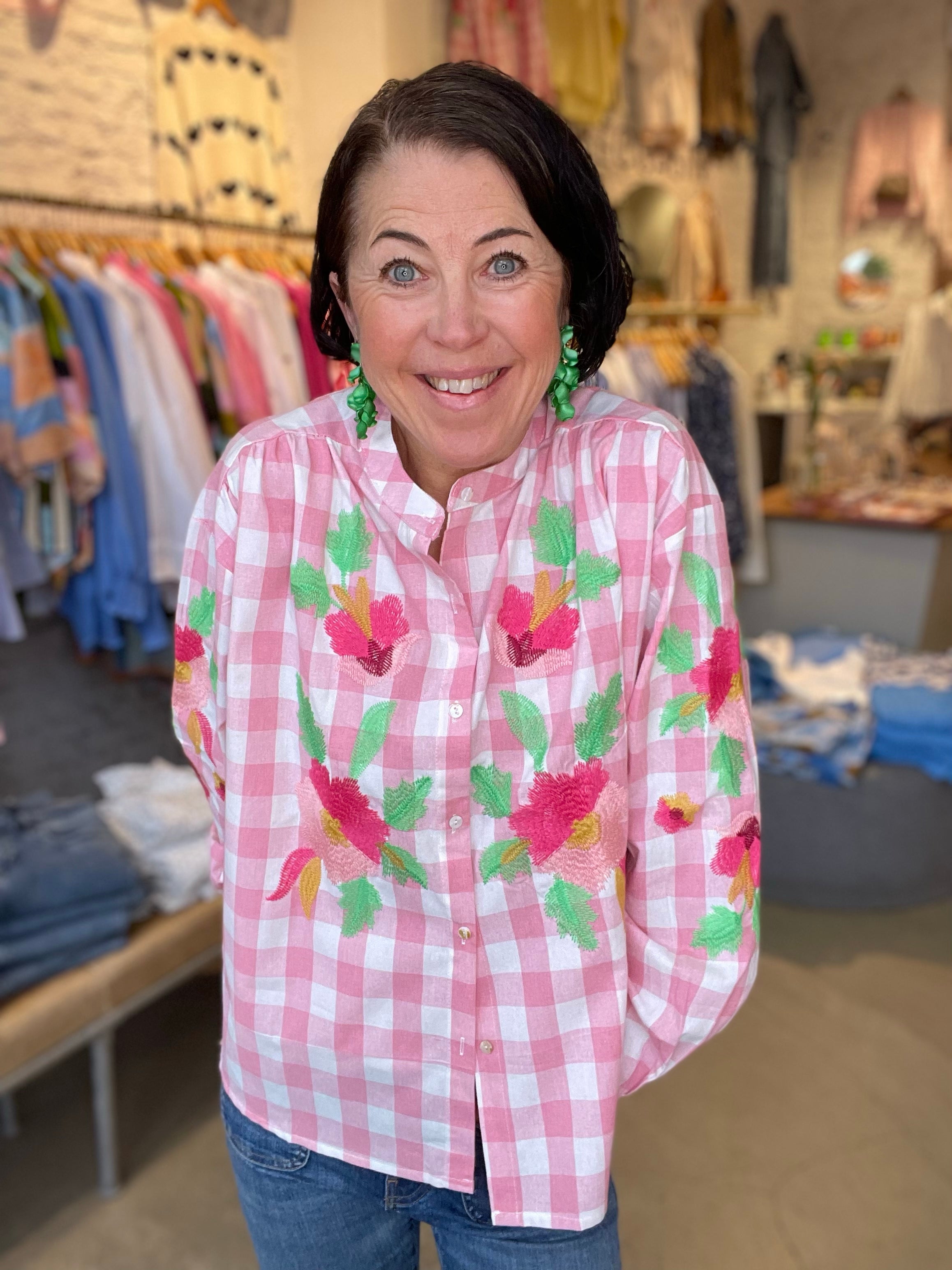 Leende kvinna framifrån i oversized rosa och vit rutig skjorta med stora broderade blommor i cerise, grönt och gult, stylade med blå jeans.