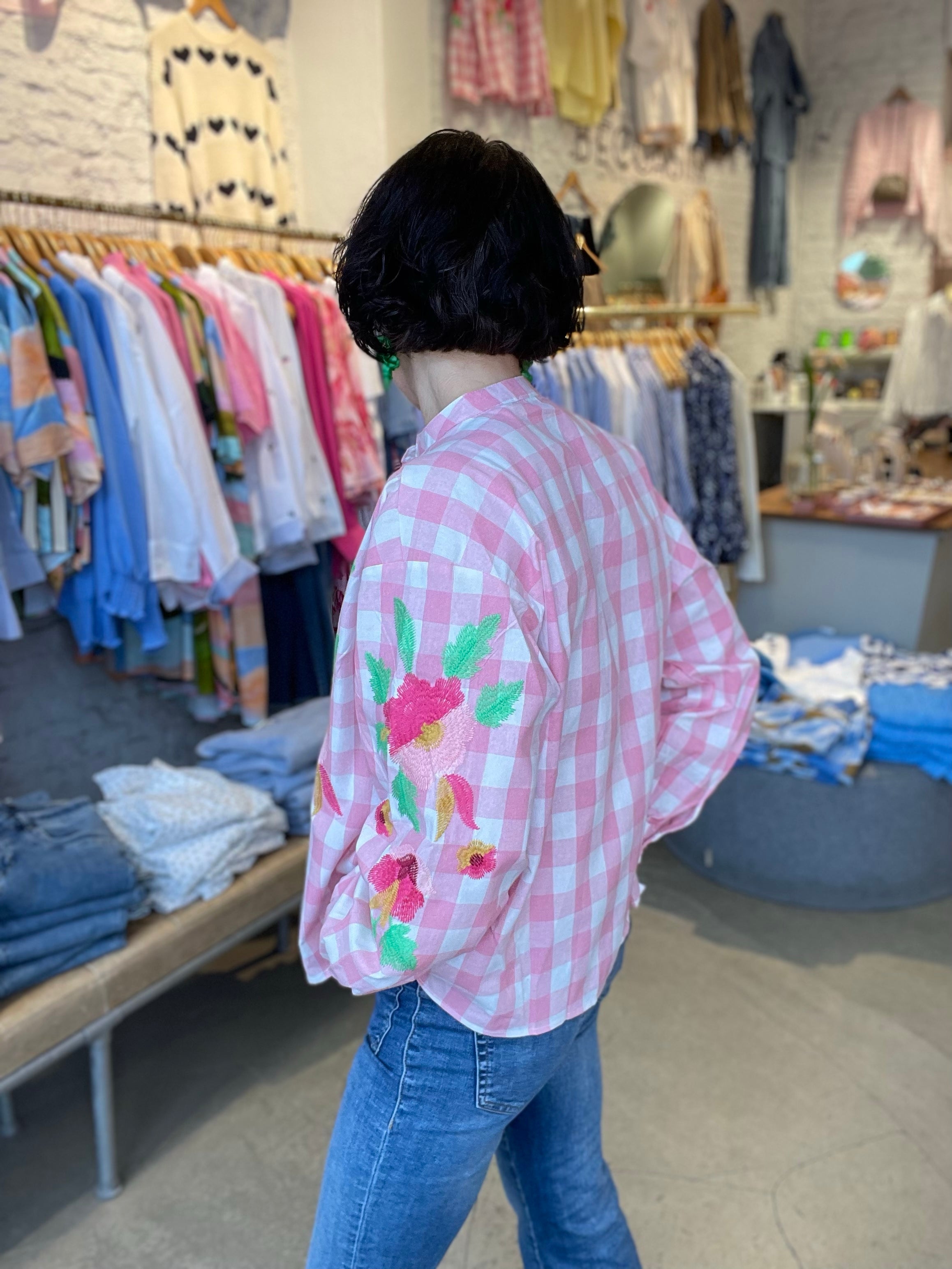 Kvinna i halvprofil bakifrån i rosa och vit rutig skjorta med broderade blommor på ärmen, stylade med blå jeans.