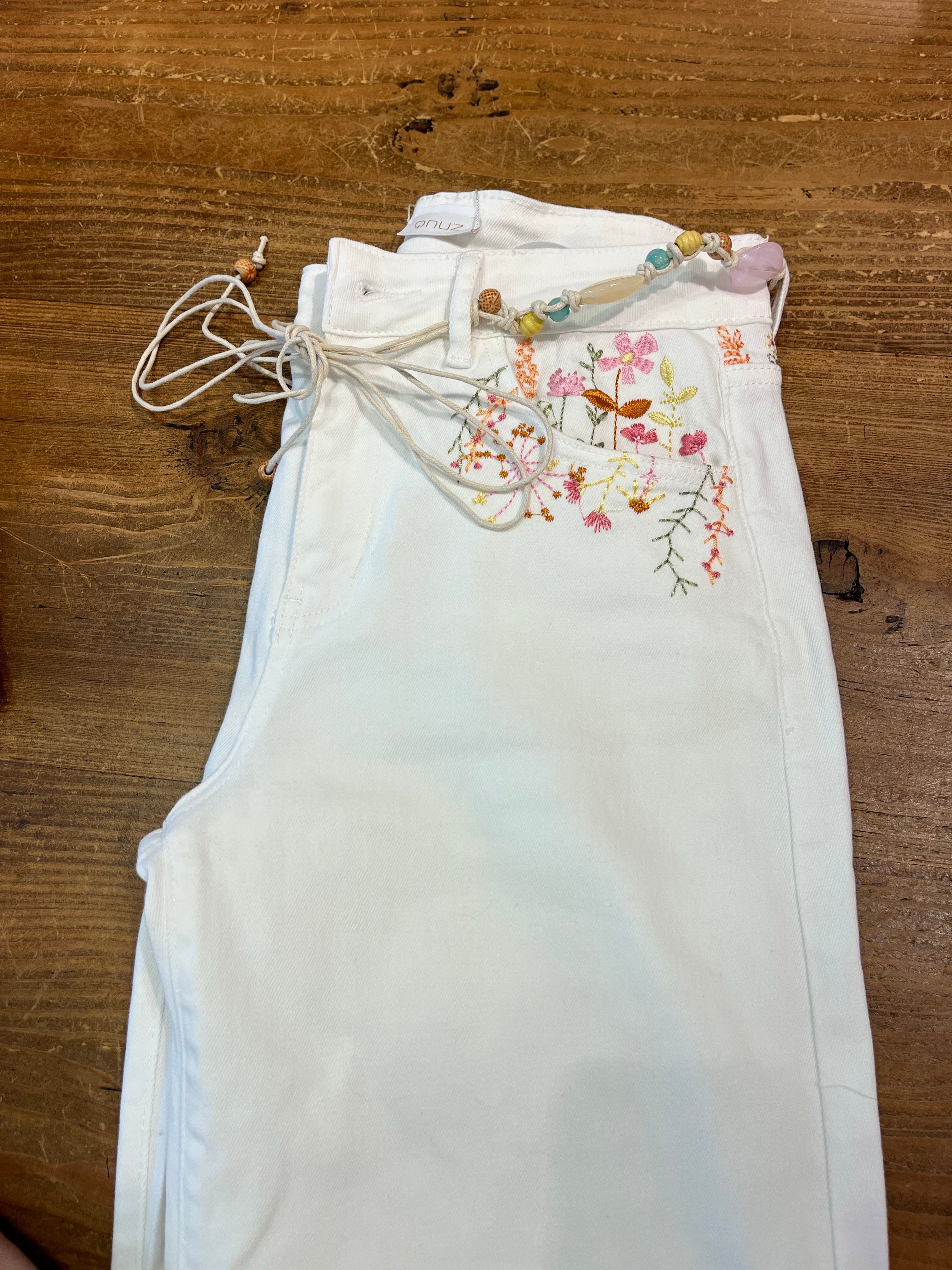 Närbild på vita jeans vikta på träbord, visar färgglatt blomsterbroderi i rosa, gult och grönt vid fickan samt knytband med pärlor och spetsdetalj längst ned.