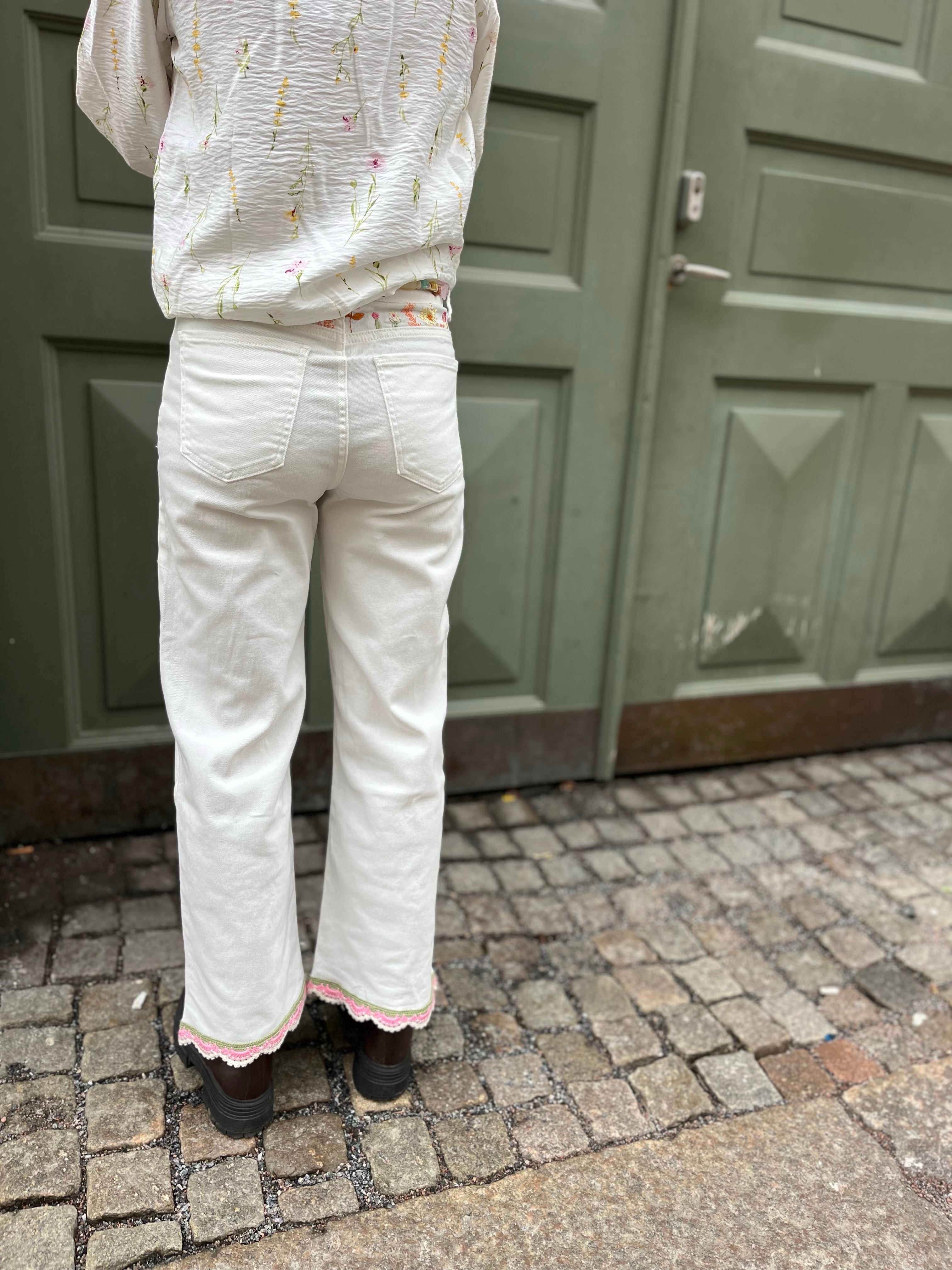 Kvinna bakifrån i vita jeans med rosa spetskant nedtill och knytband i midjan, stylade med vit blommig skjorta och bruna boots.