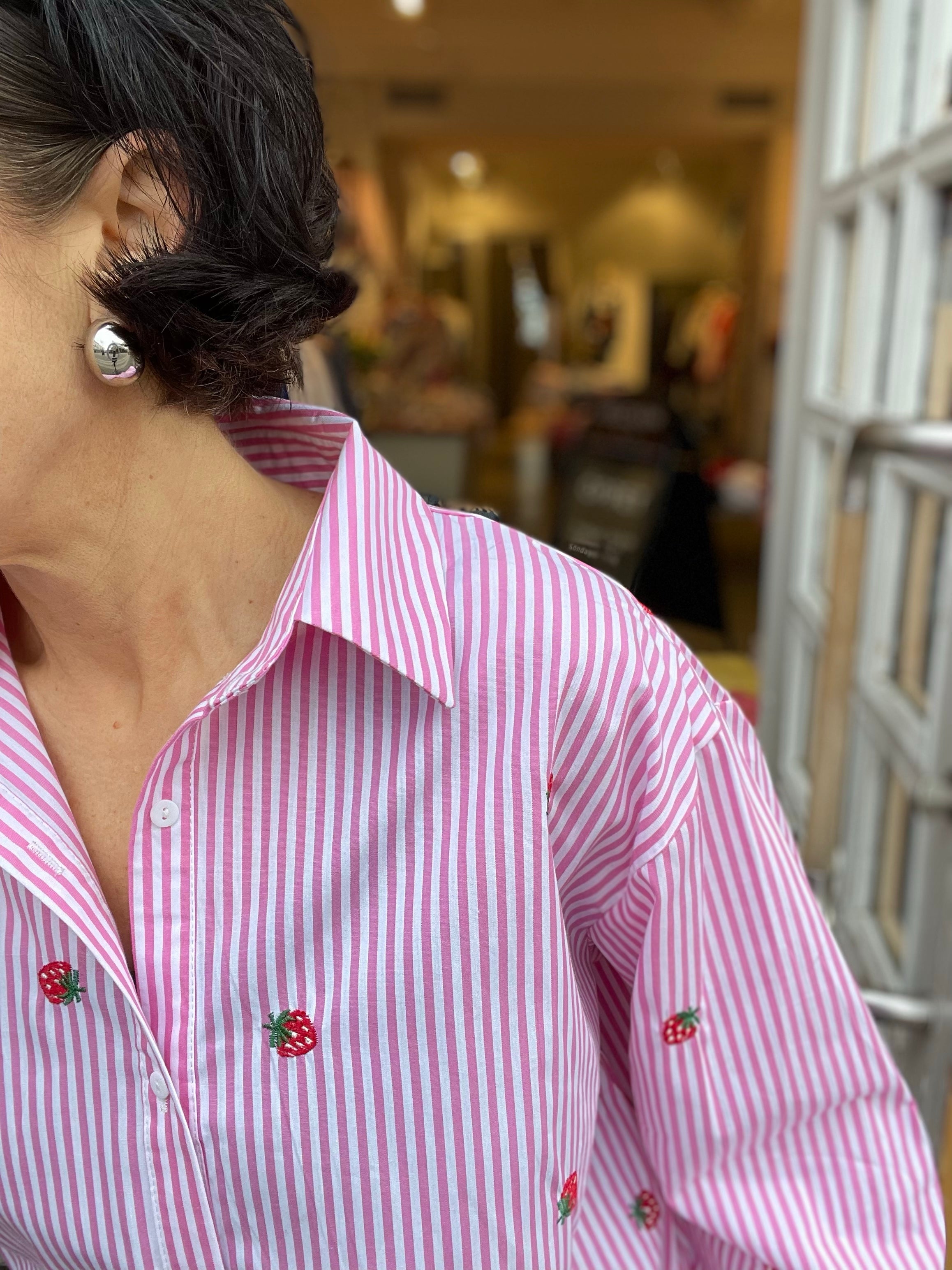 Närbild på krage och överkropp av rosa randig skjorta med broderade jordgubbar och klassisk krage, kvinna med silverrunda örhängen fotograferad i butiksdörren.