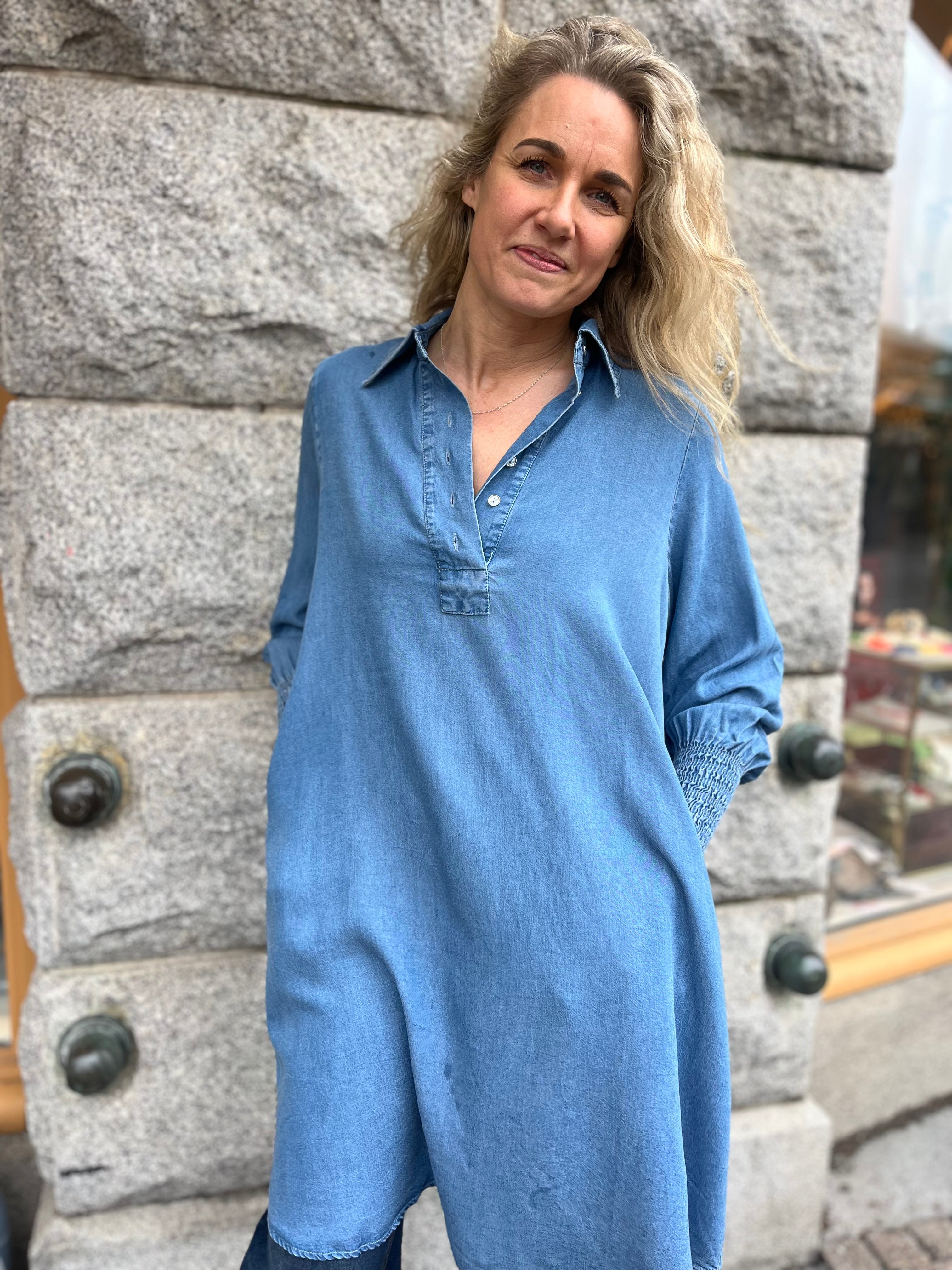 Helbild av kvinna i oversized denimblå skjortklänning med klassisk krage och smockade ärmslut, stylade med blå jeans – utomhus mot stenvägg.