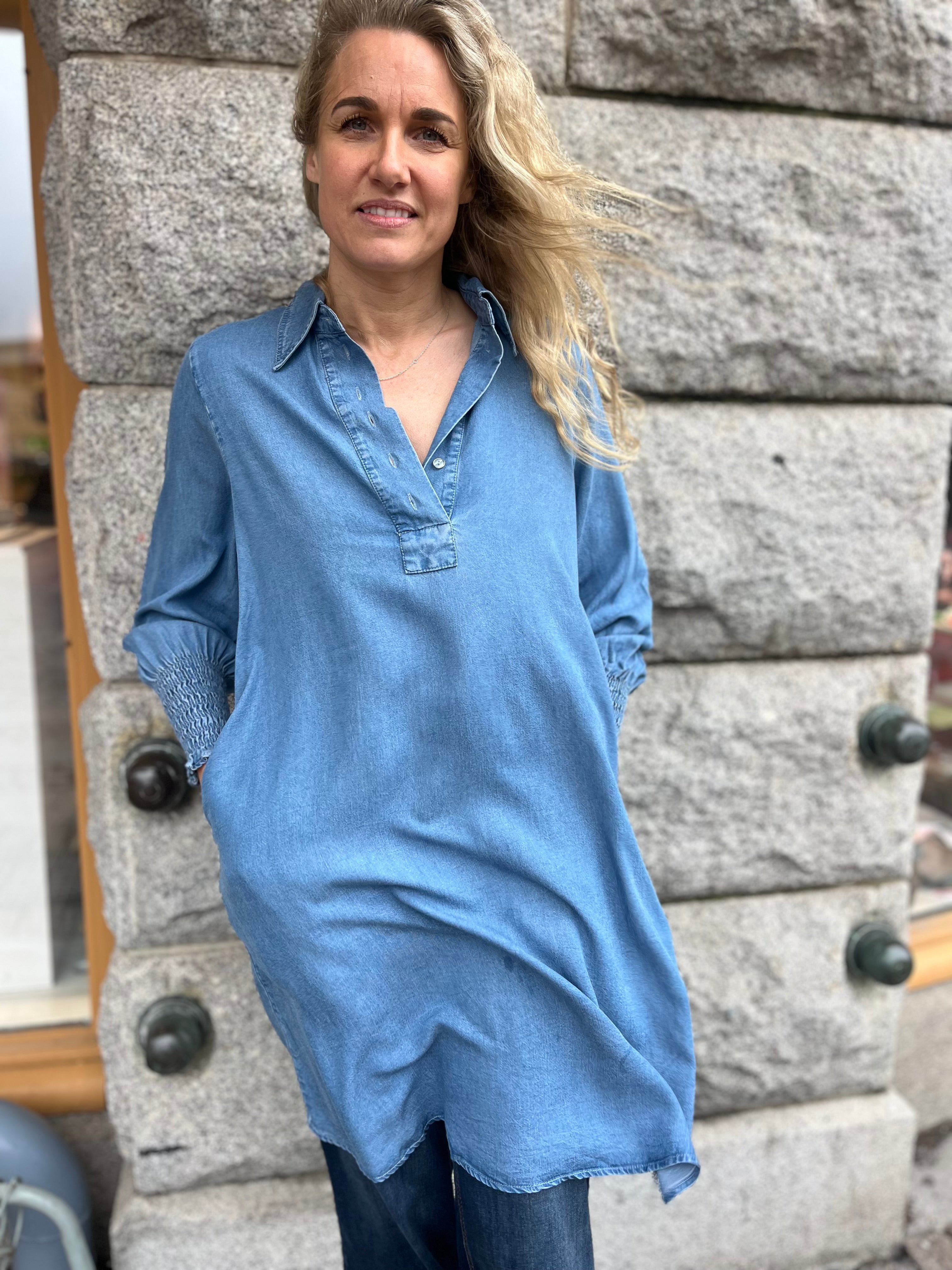 Kvinna i lång denimblå skjortklänning med V-ringning, klassisk krage, halvknäppning och smockade ärmslut – fotograferad utomhus mot grå stenvägg.