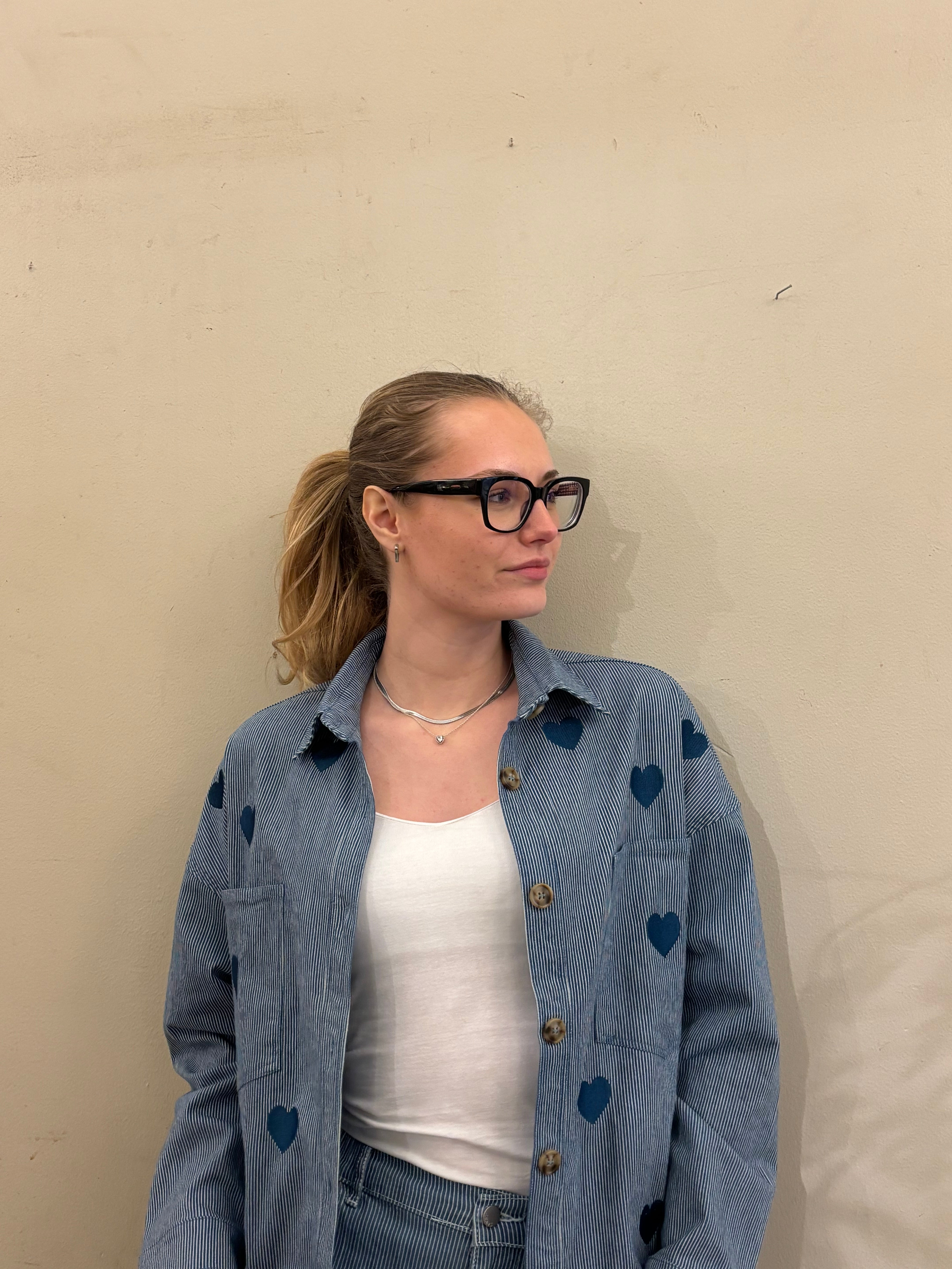 Kvinna i blåvitrandig skjortjacka med hjärtmönster, öppen över vit linne – närbild överkropp mot beige vägg.