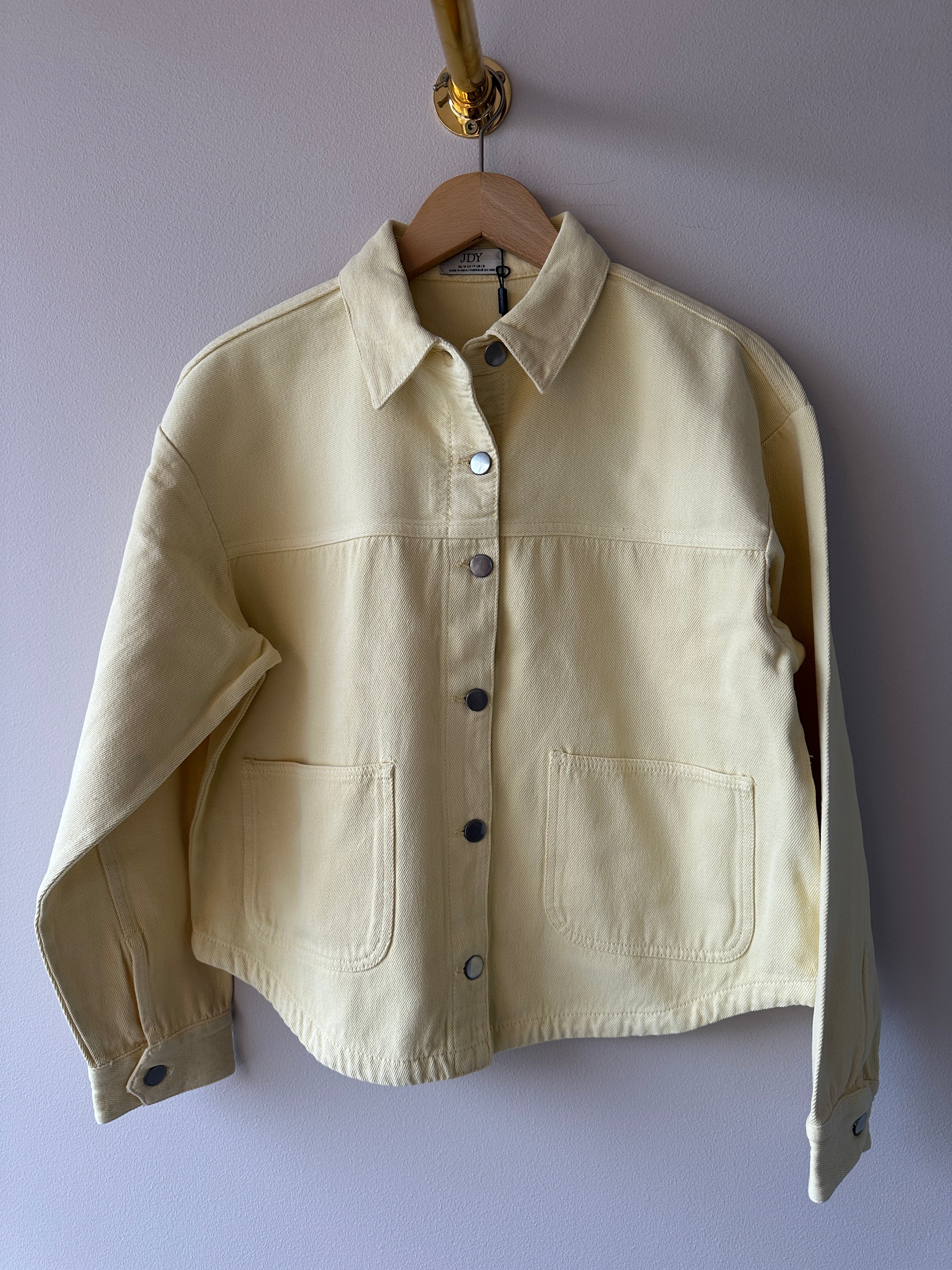 Crèmefärgad oversized shacket i denimtyg med klassisk krage, tryckknappar och stora patchfickor, hängd på galge mot ljuslila vägg.