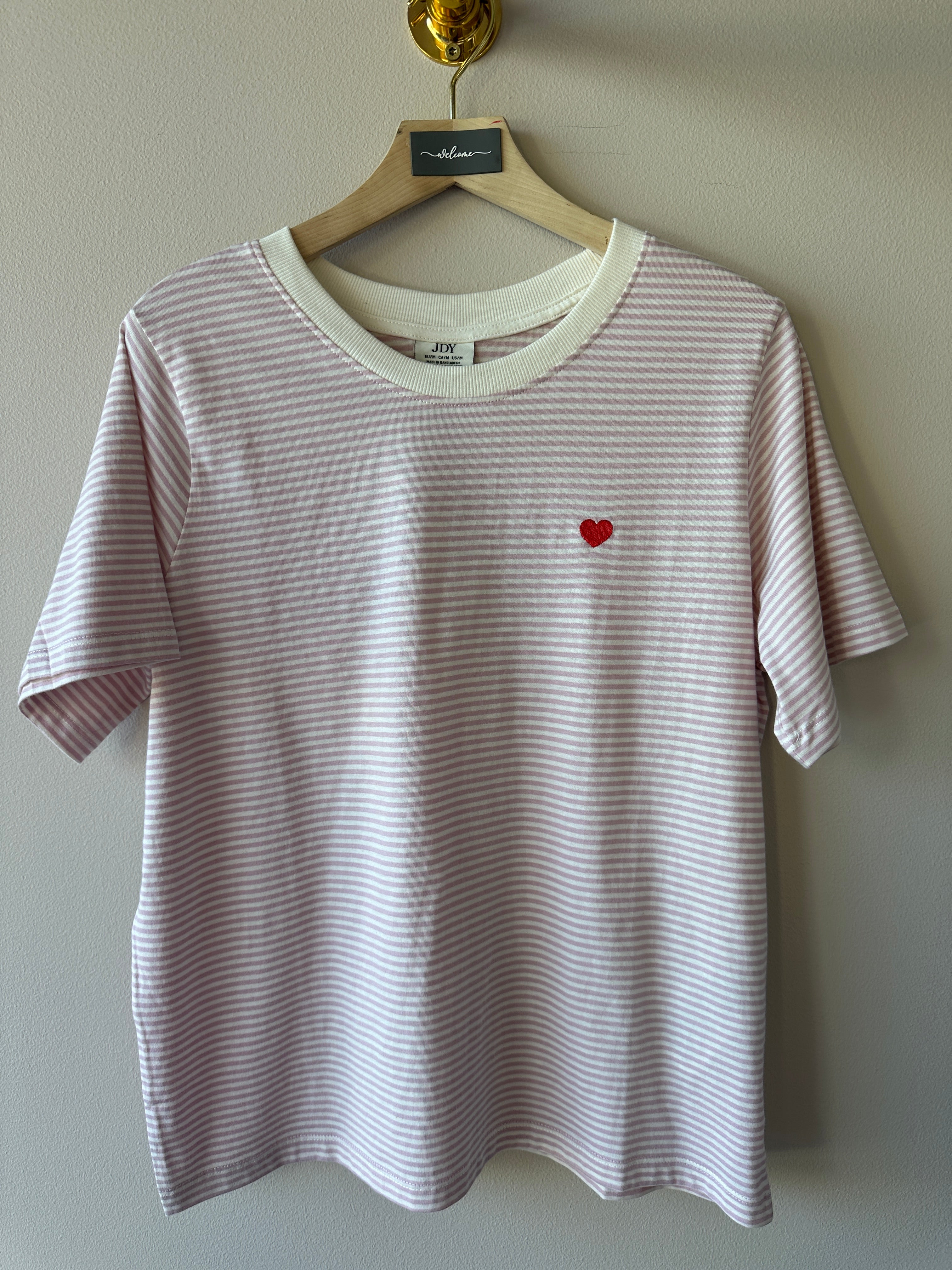 Lila och vit randig ribbad t-shirt med litet rött hjärtbroderi på bröstet, rund hals och korta ärmar hängande på galge mot beige vägg.