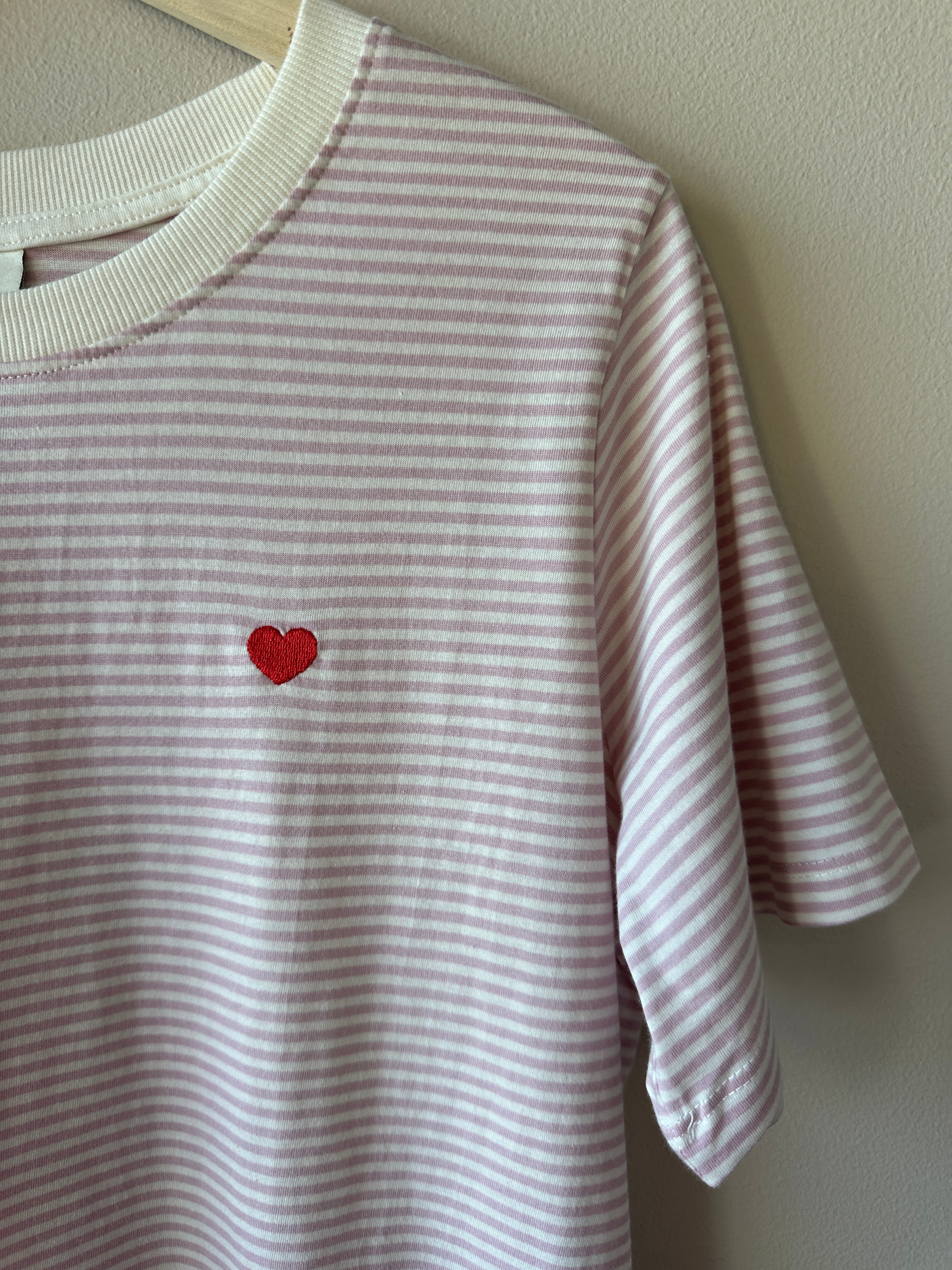 Närbild på lila och vit randig ribbad t-shirt som visar litet rött hjärtbroderi på bröstet och ribbad rund hals, hängande på galge.