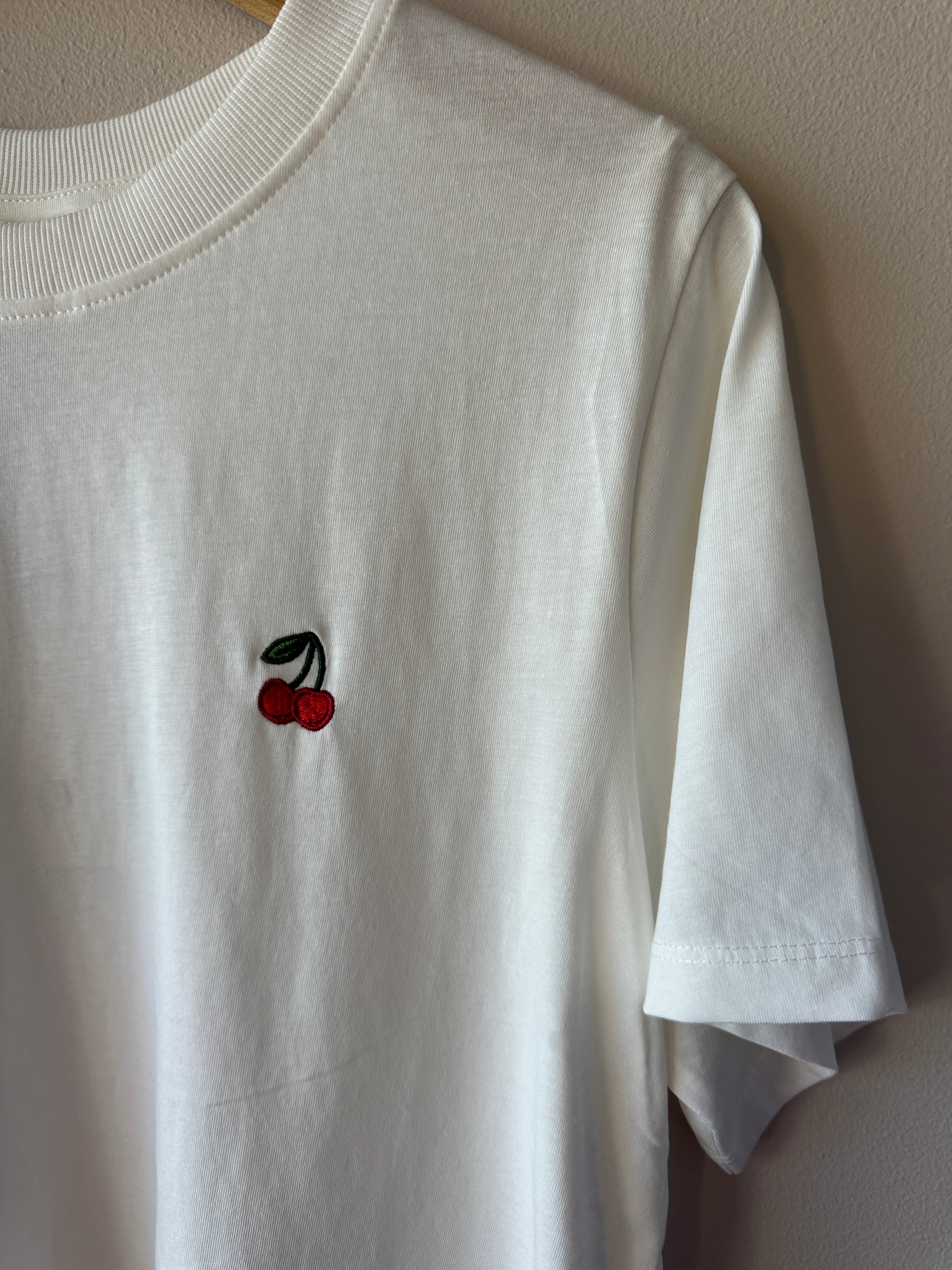 Närbild på vit t-shirt som visar litet rött körsbroderi med gröna blad på bröstet och ribbad rund hals, hängande på galge.
