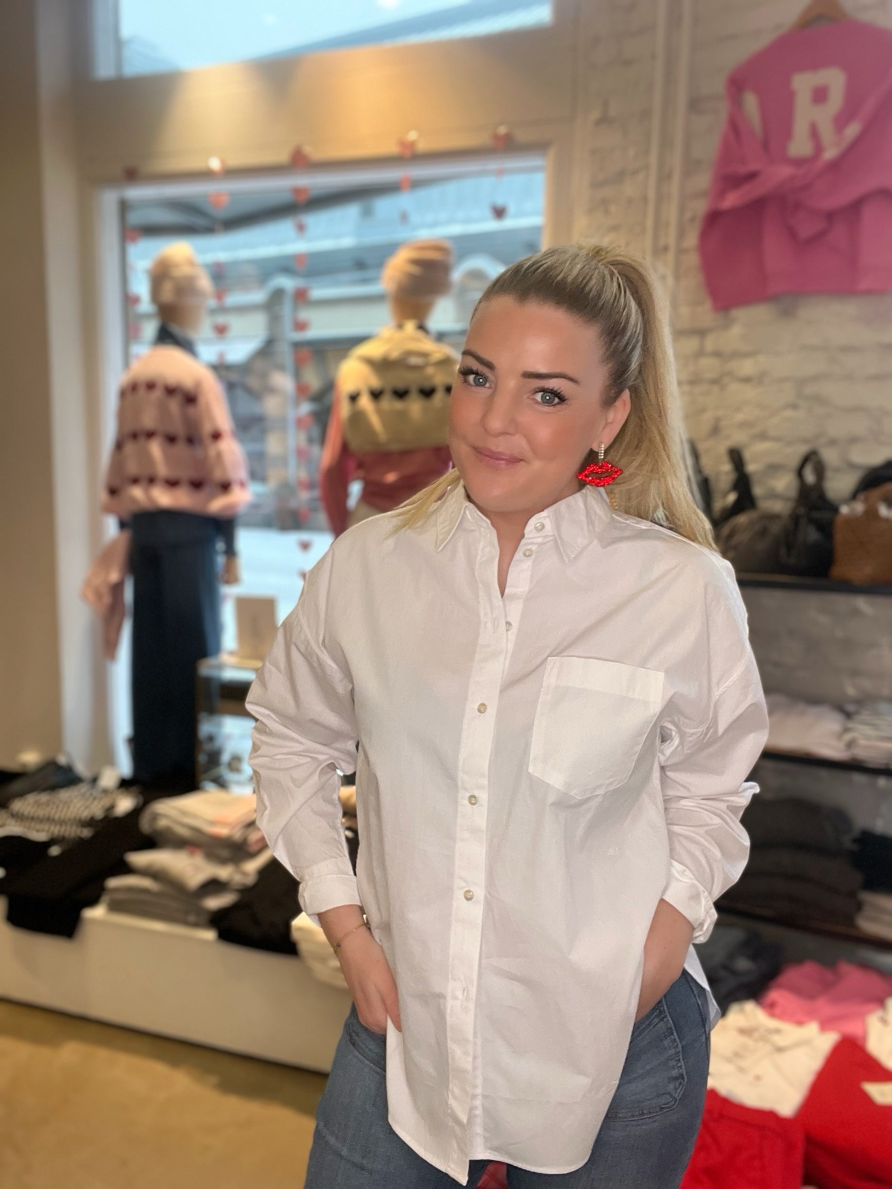 Kvinna framifrån med händerna i fickorna i oversized vit poplinskjorta med bröstficka och klassisk krage, stylade med blå jeans och röda läppörhängen, fotograferad i butiksmiljö.