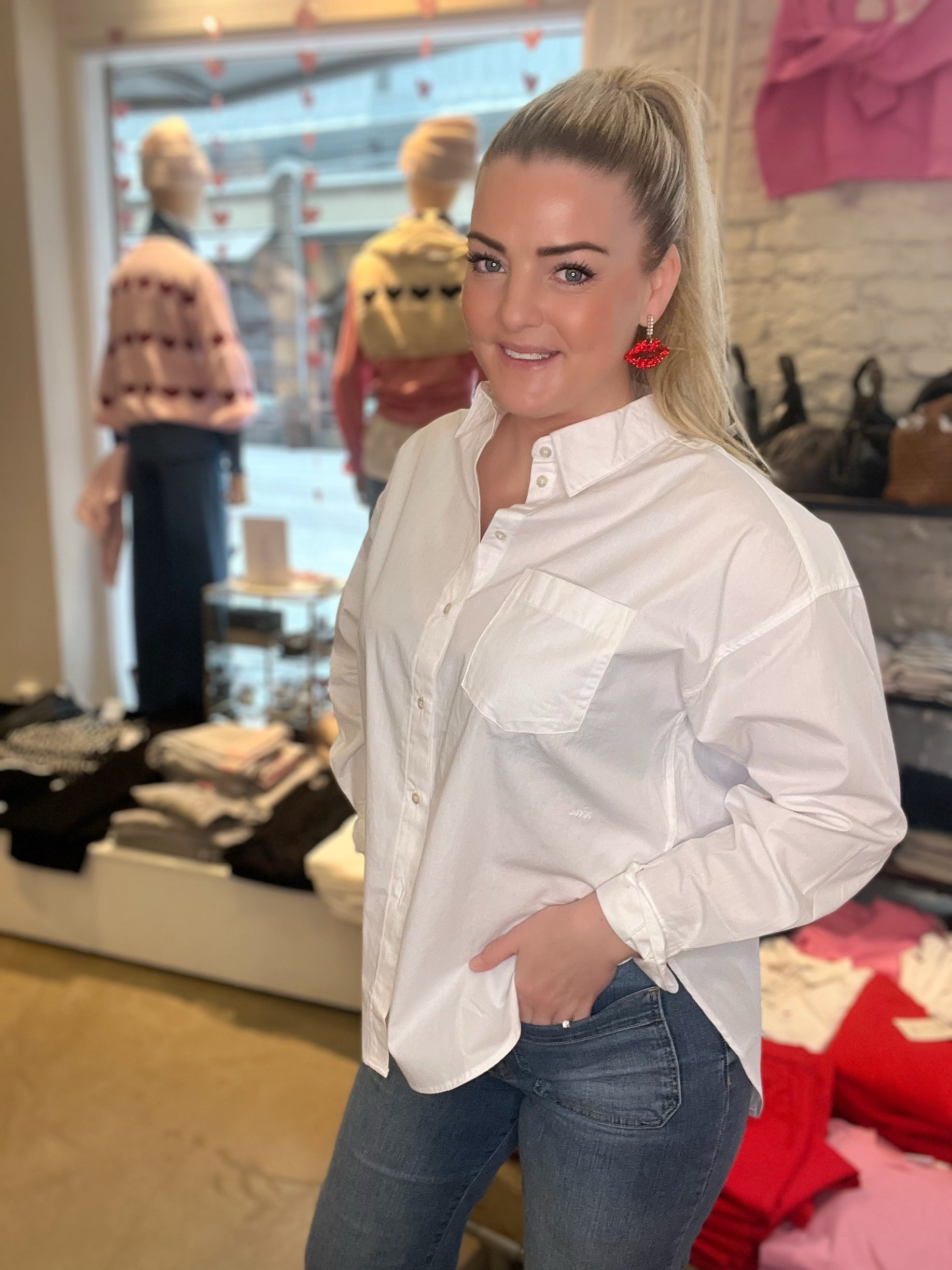 Leende kvinna i halvprofil i oversized vit poplinskjorta med bröstficka och handen i fickan, stylade med blå jeans och röda läppörhängen, fotograferad i butiksmiljö med skyltfönster i bakgrunden.
