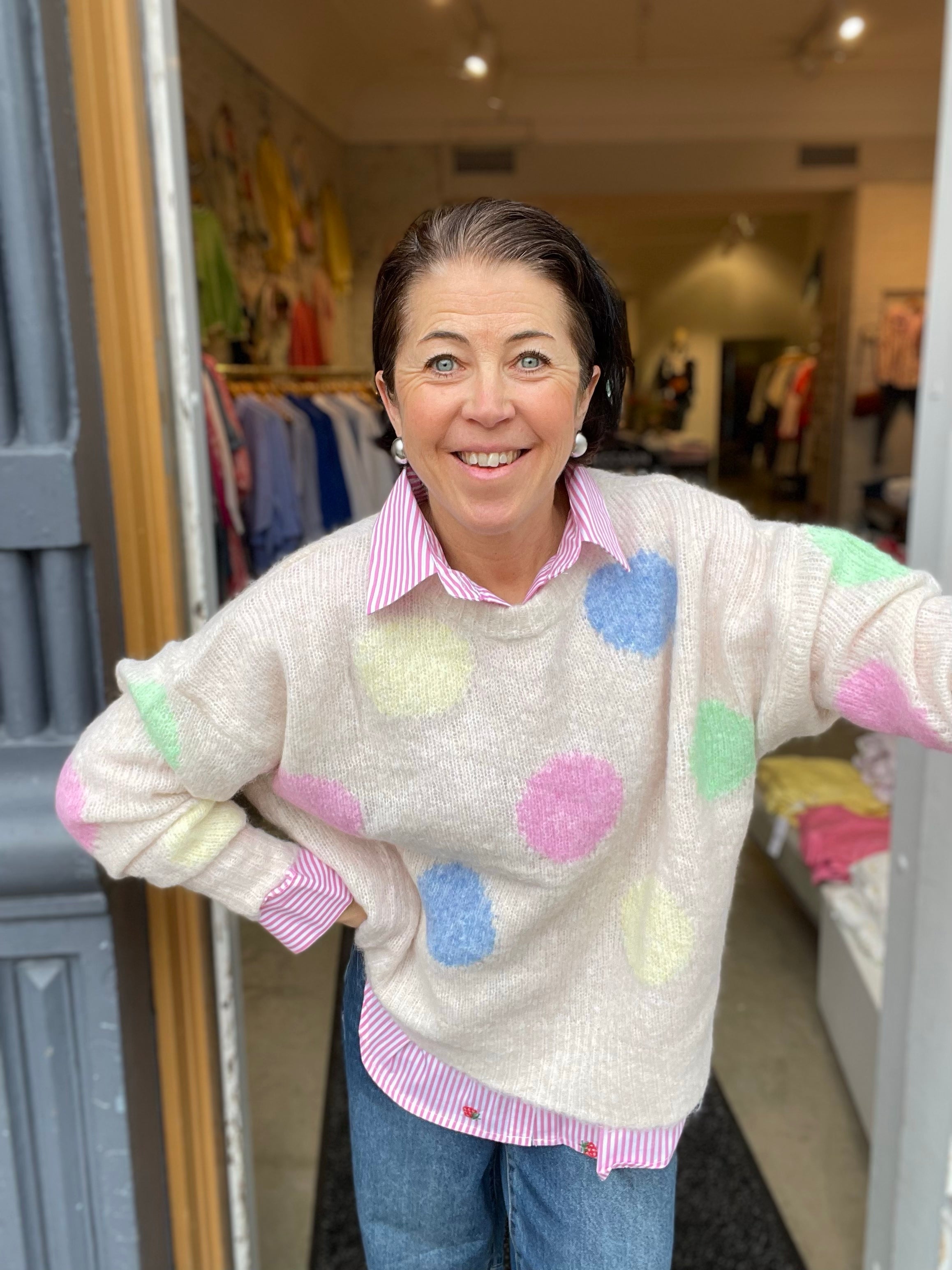 Leende kvinna med händerna i sidorna i oversized ecru stickad tröja med stora pastellfärgade prickar i rosa, blått, grönt och gult, stylade med randig rosa skjorta och blå jeans.