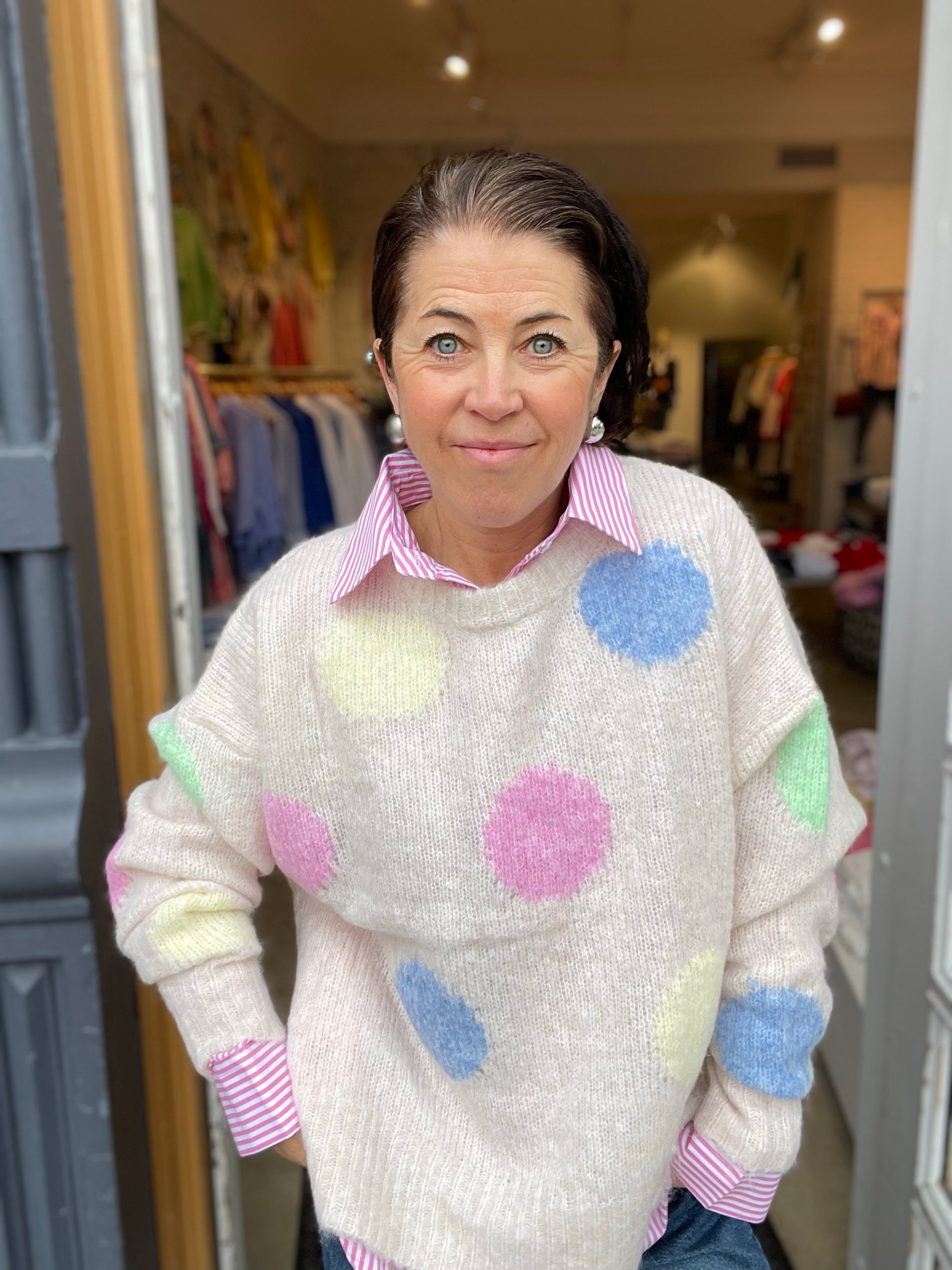 Närbild på kvinna i ecru stickad tröja med stora fluffiga pastellprickar och randig rosa skjorta under, leende mot kameran i butiksdörren.