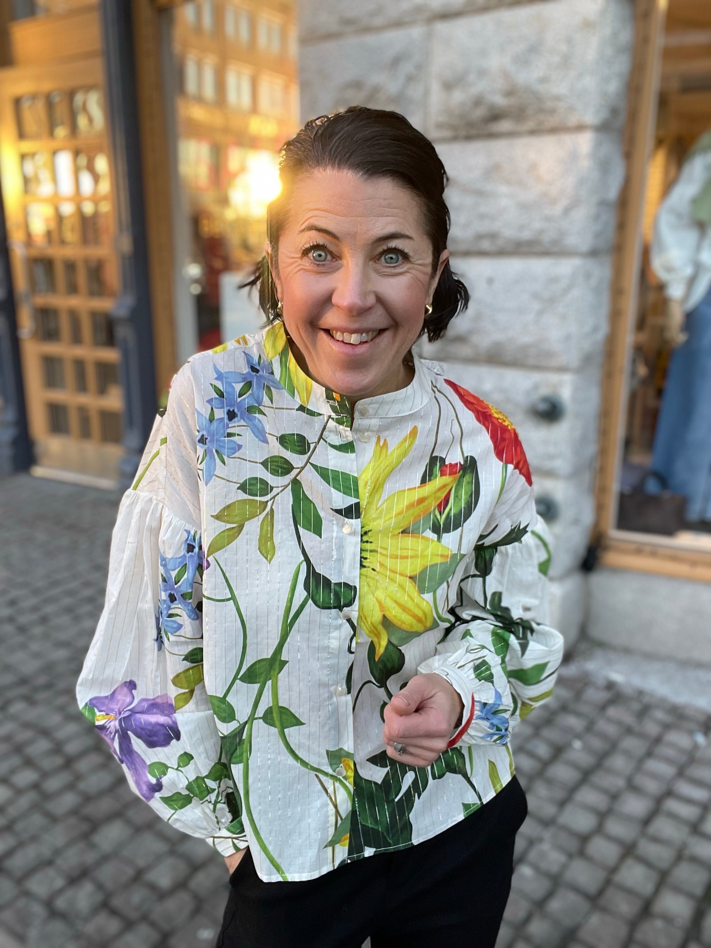 Kvinna närbild i vit skjorta med stort färgglatt blomtryck i gult, rött, lila och blått, stående krage och ballongärmar, utomhus i kvällsljus.