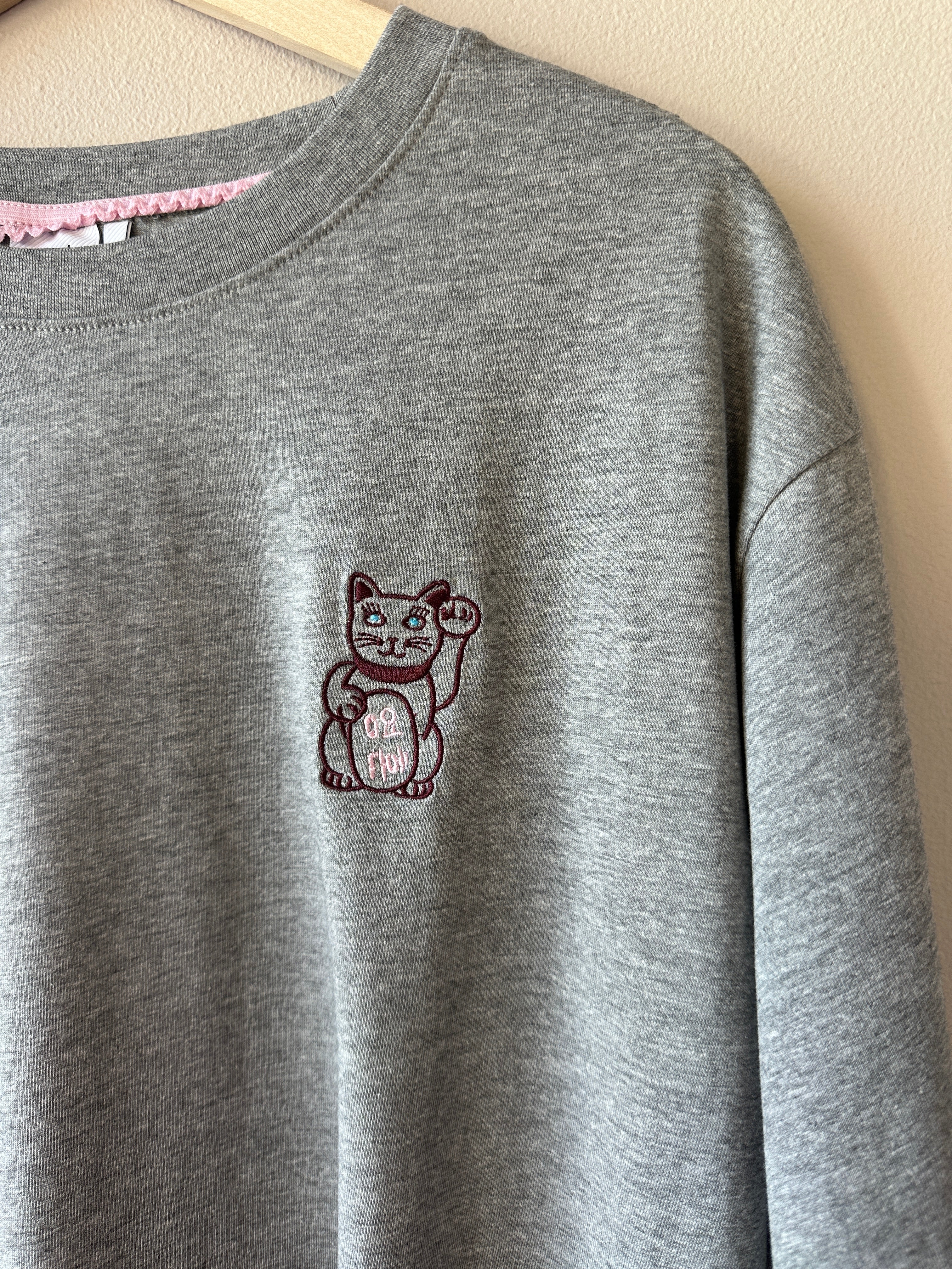 Närbild på bröstet av gråmelerad t-shirt som visar detaljerat broderi av en japansk lyckokatt med lyft tass i mörkrött och rosa mot grått tyg.