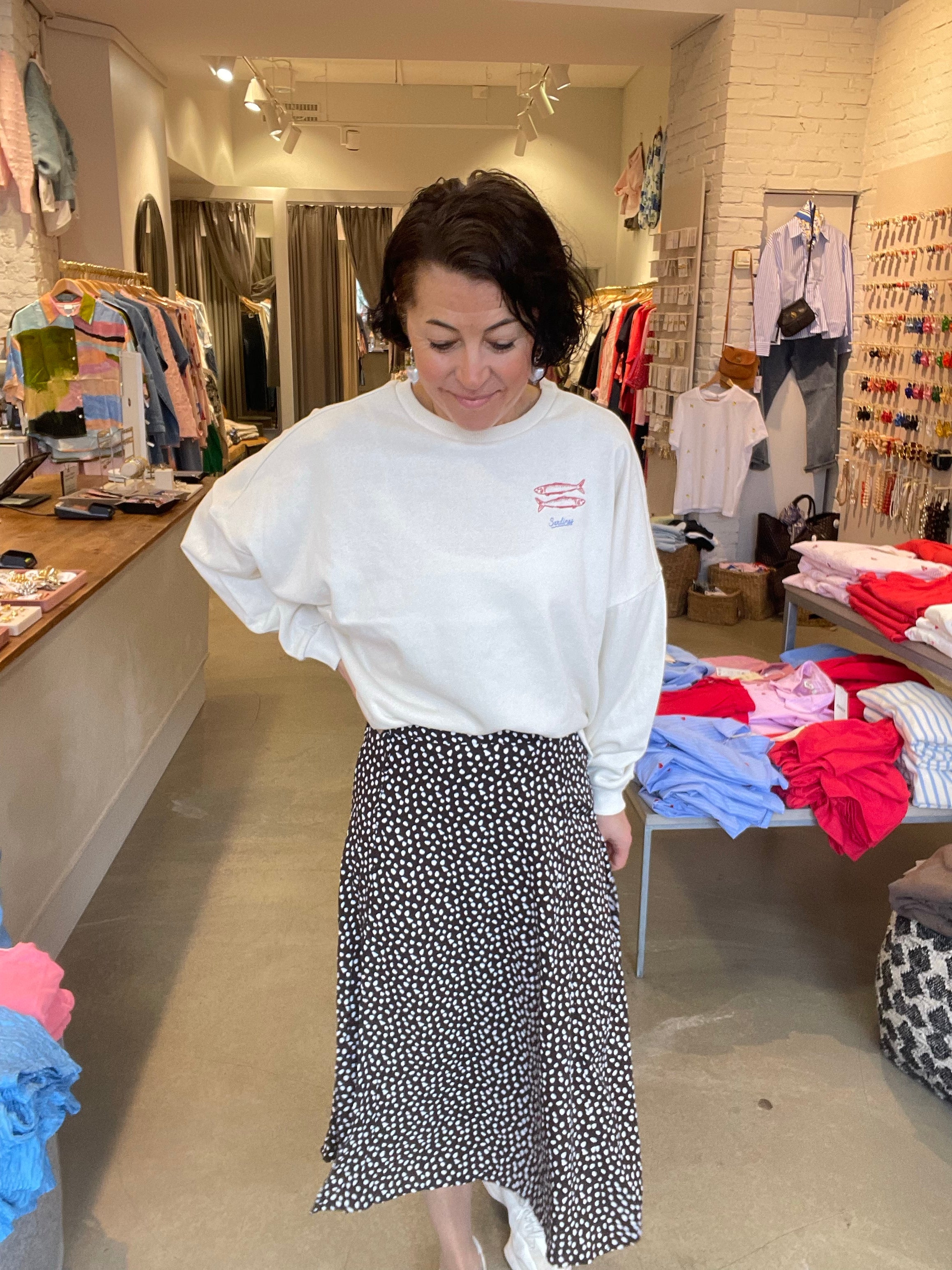 Kvinna framifrån i svart prickig midikjol med vit sweatshirt och handen sidan, fotograferad i butiksmiljö med klädstativ i bakgrunden.