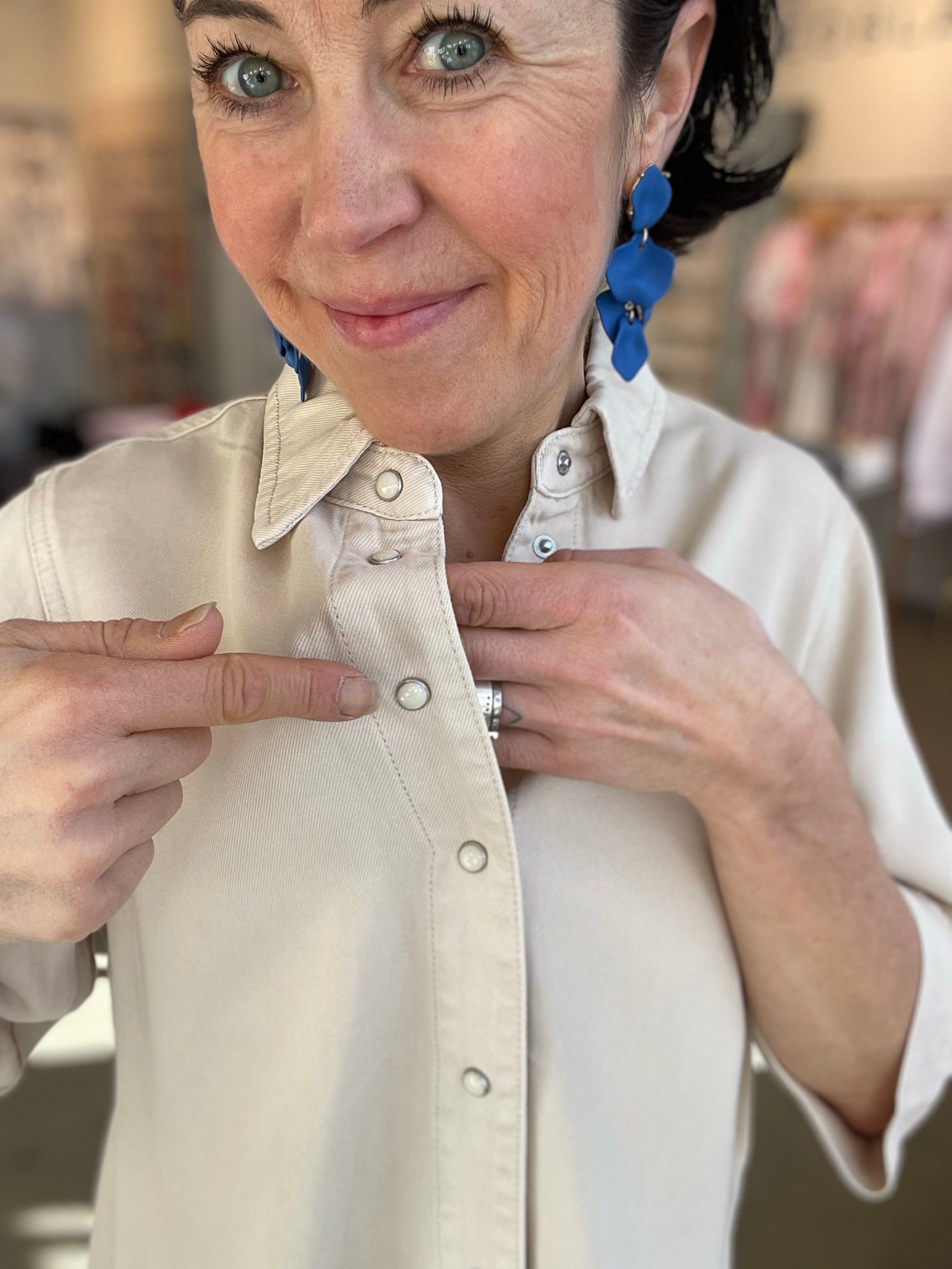 Kvinna närbild som visar tryckknappar i silver på beige denimtopp med klassisk krage, stylade med blå blommörhängen och ring, inomhus i butik.