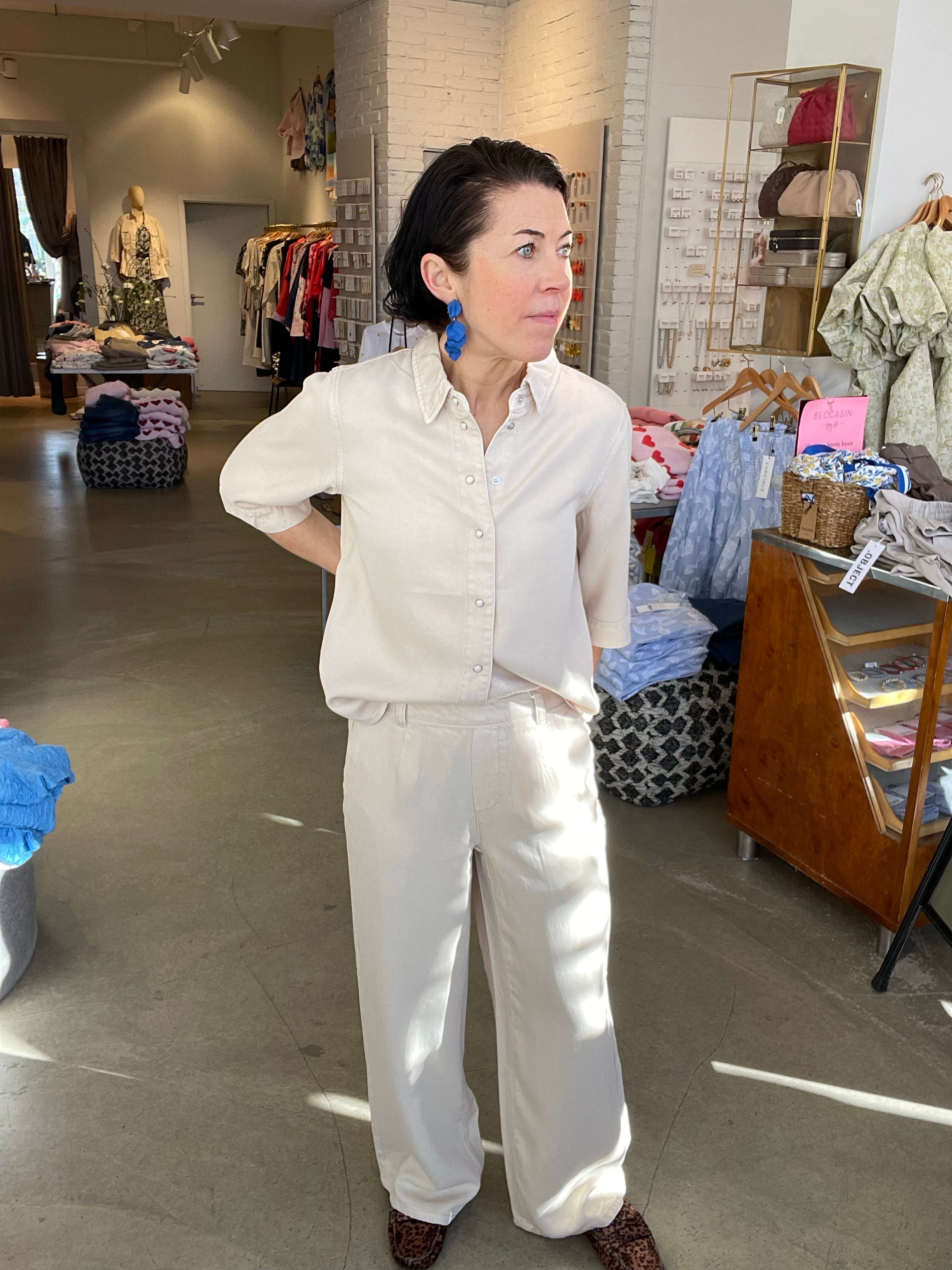 Kvinna framifrån med handen i sidan i beige wide leg-byxor med hög midja, stylade med matchande beige denimtopp, blå blommörhängen och leopardmönstrade skor, inomhus i butik.