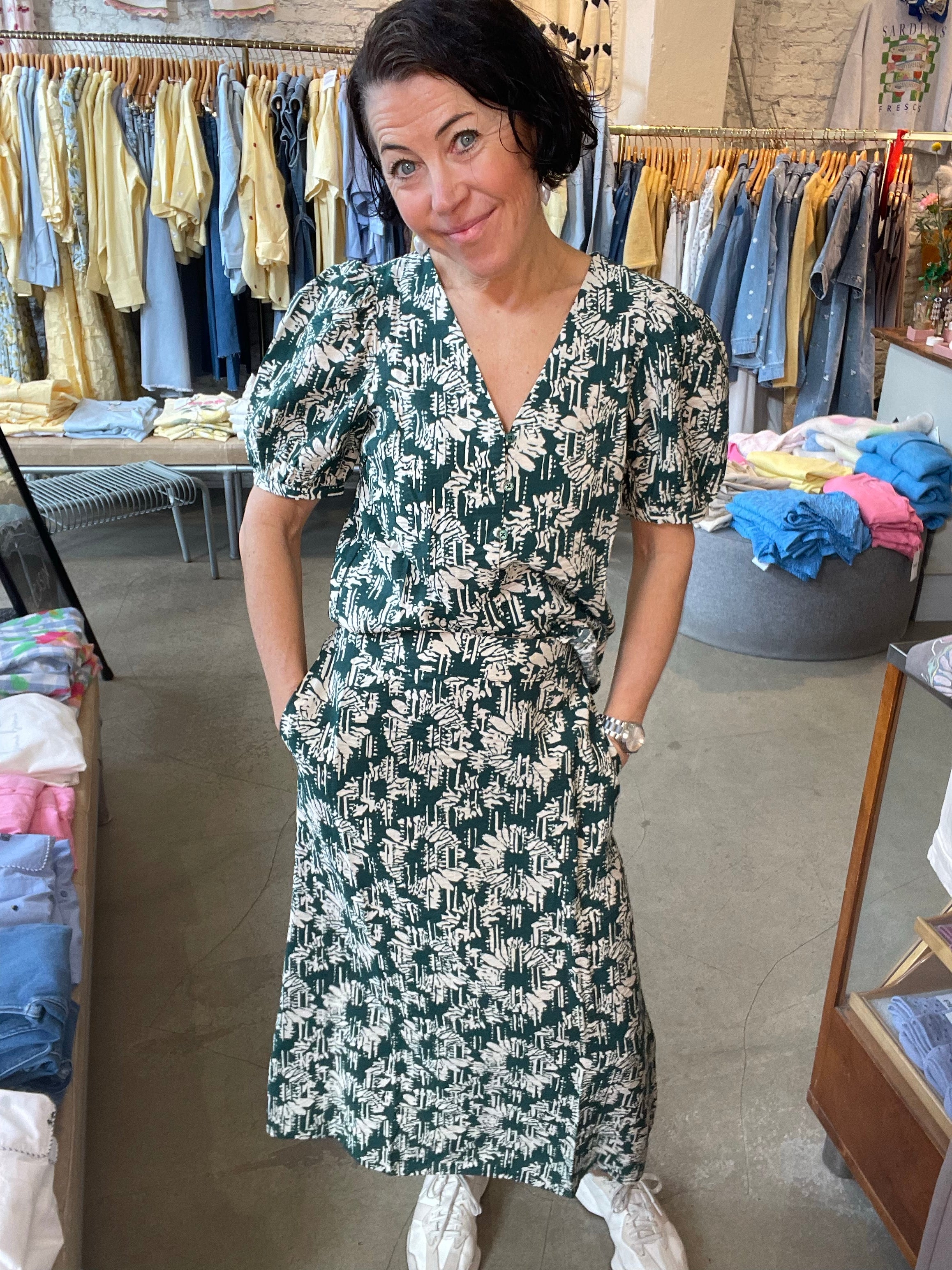 Leende kvinna framifrån i mörkgrön och beige abstrakt midikjol med matchande topp, händerna i fickorna och vita sneakers, fotograferad i butiksmiljö.