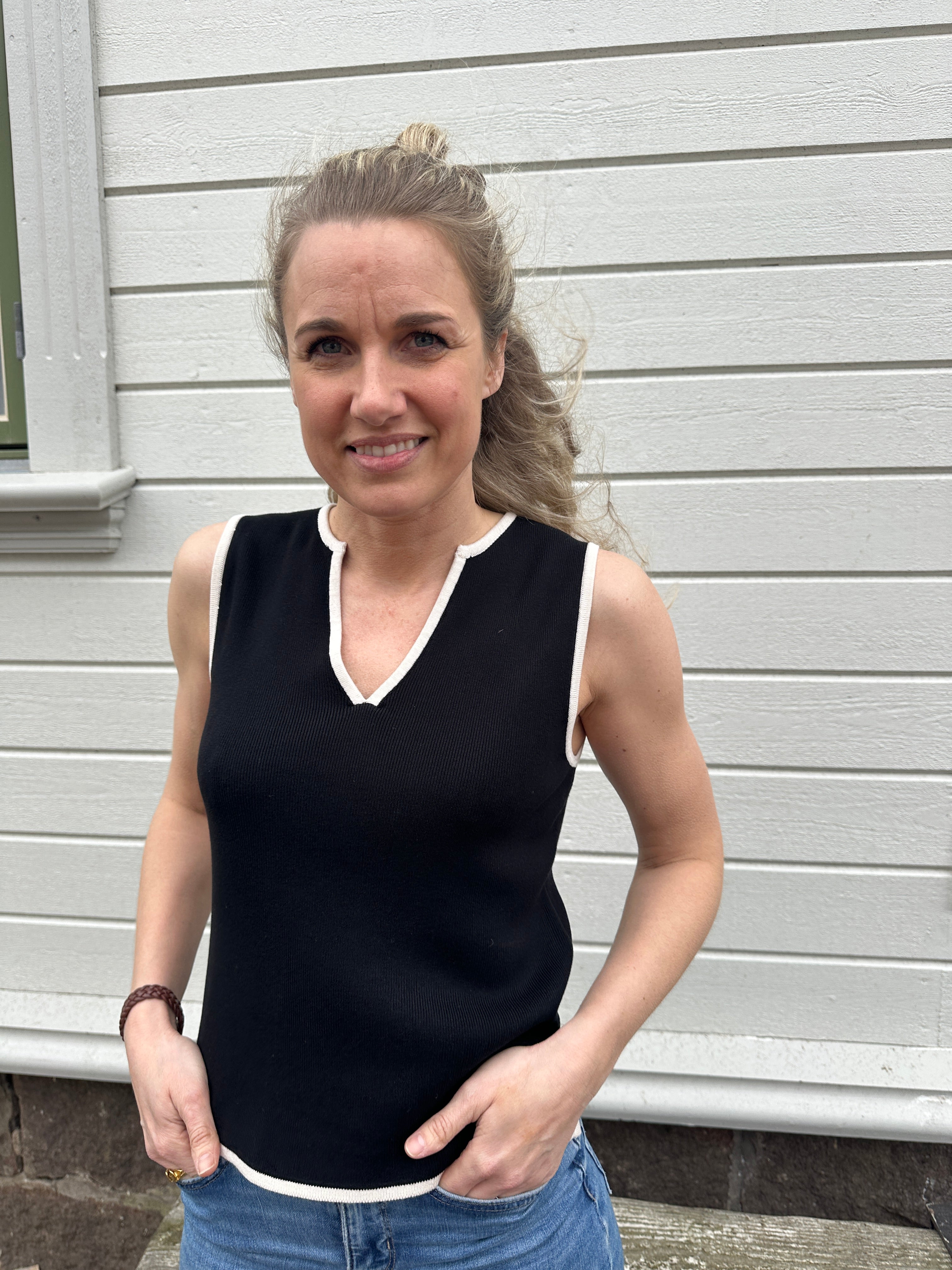 Kvinna iklädd svart ärmlös stickad topp med v-ringning och vita kontrastkanter vid hals och ärmhål, poserar med händerna i fickorna framför vit träfasad.