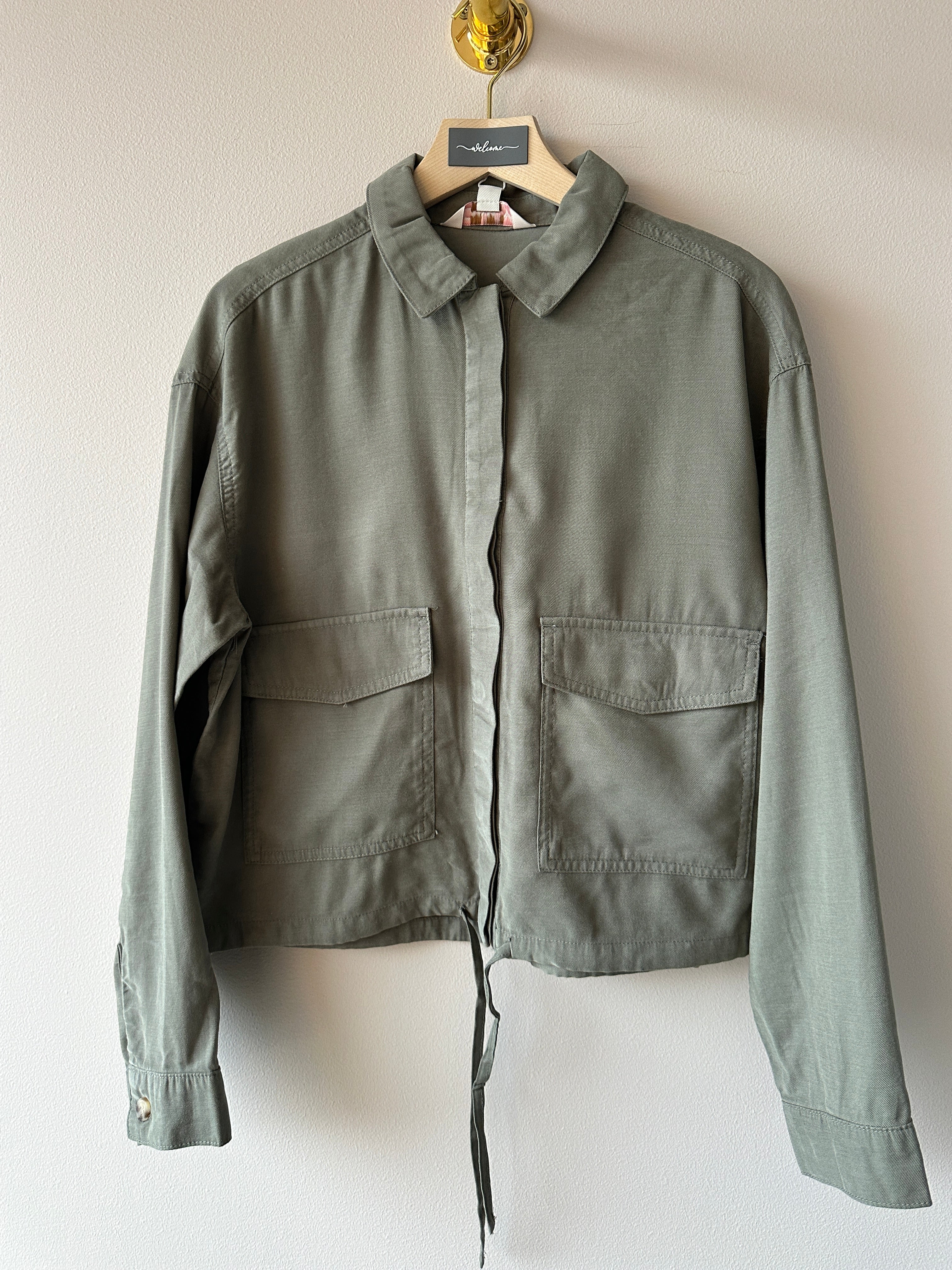 Olivgrön skjortjacka med klassisk krage, dolda knappar, stora brystfickor och knytband nedtill, hängd på galge mot ljus vägg.
