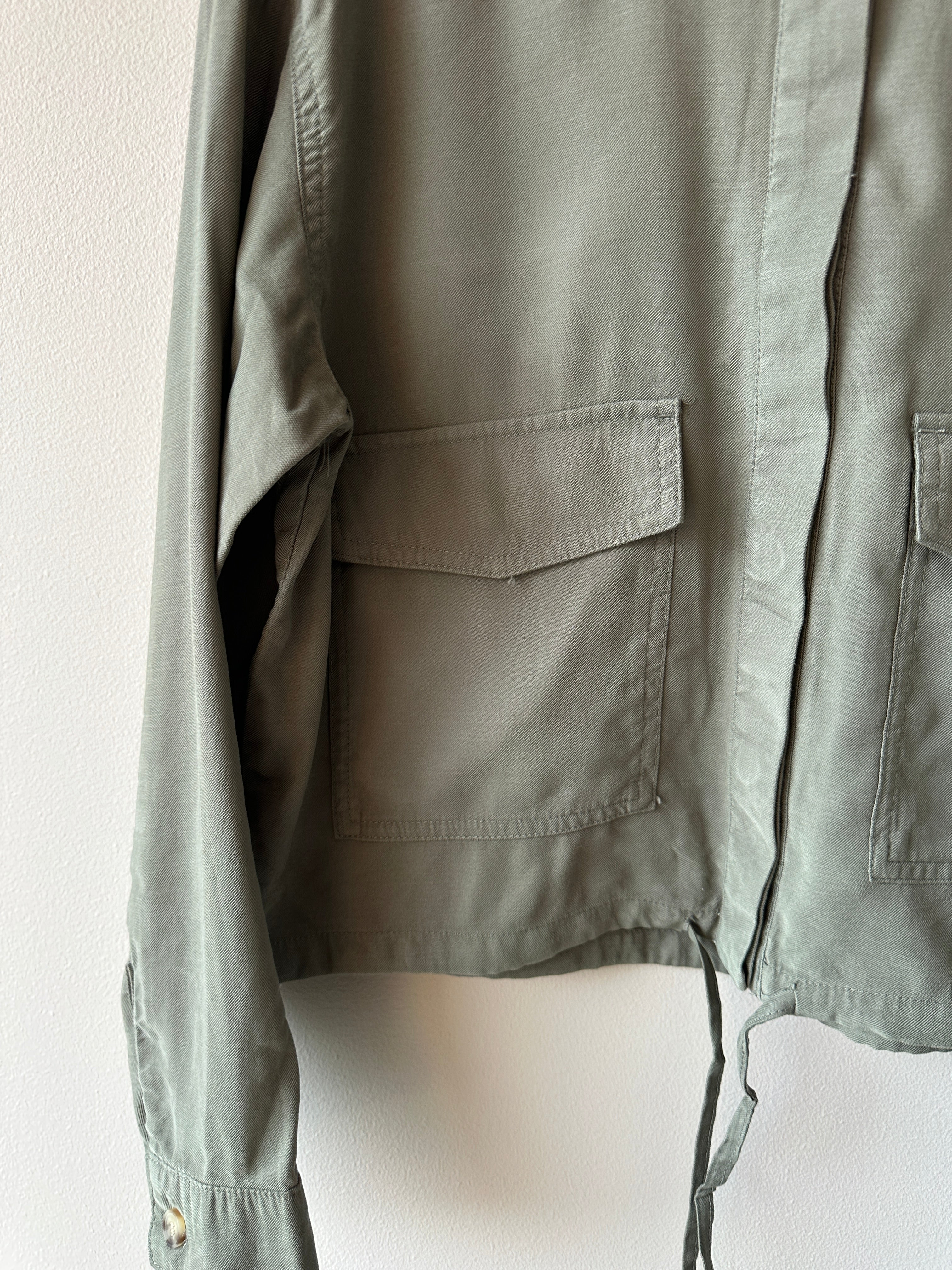 Närbild på sidan av olivgrön skjortjacka med stor patchficka, knytband i midjan och tryckknappar vid ärmen.