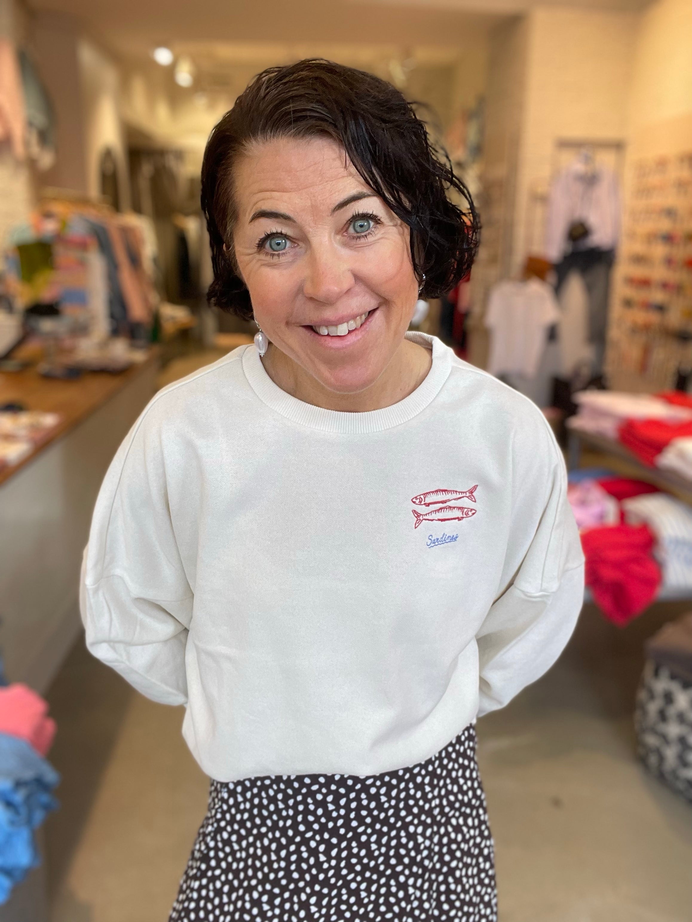 Leende kvinna framifrån i oversized vit sweatshirt med litet broderat sardinmotiv på bröstet, stylade med svart prickig kjol, fotograferad i butiksmiljö.