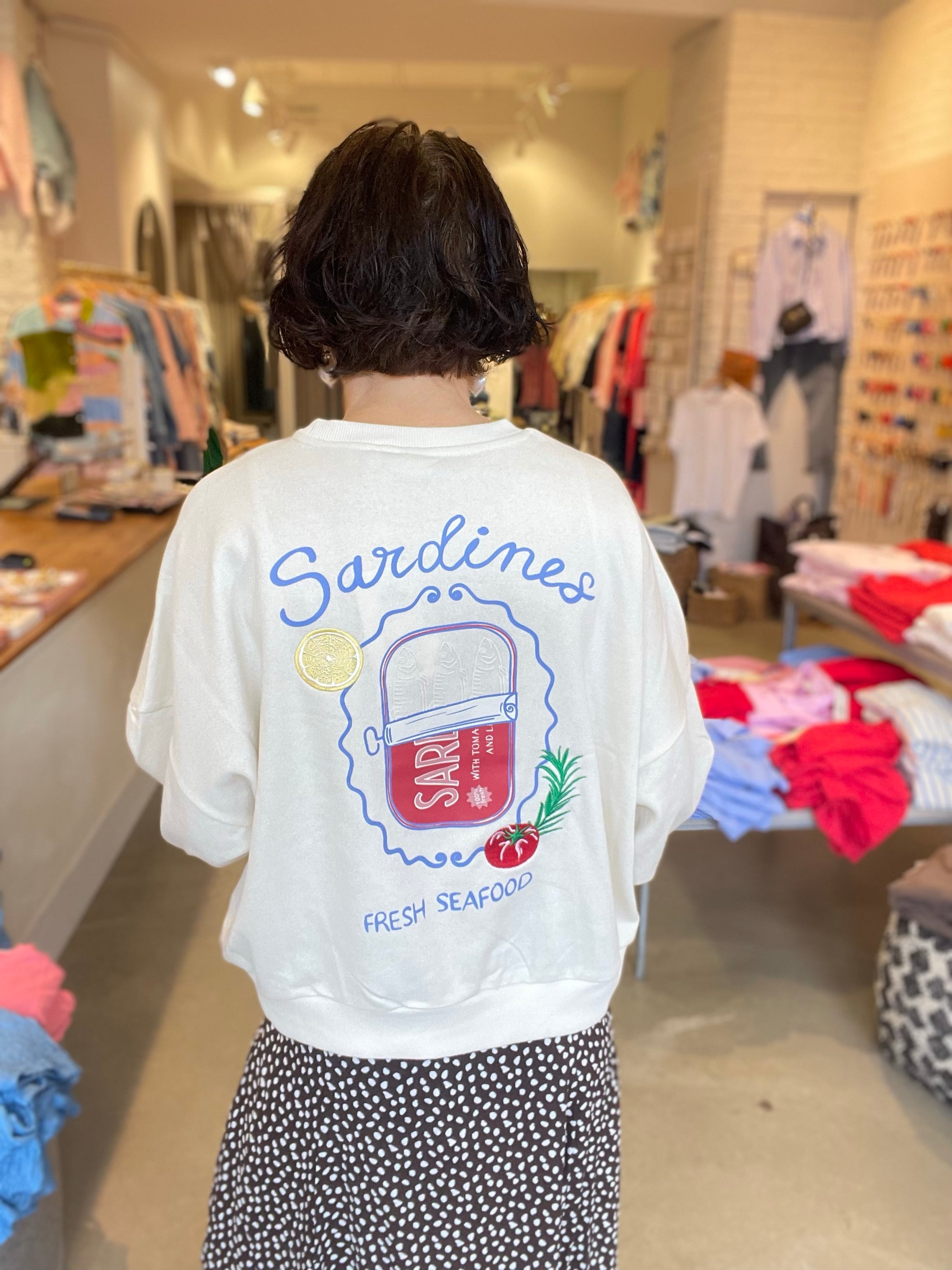 Kvinna bakifrån i vit sweatshirt med stort grafiskt Sardines Fresh Seafood-tryck på ryggen, stylade med svart prickig kjol.