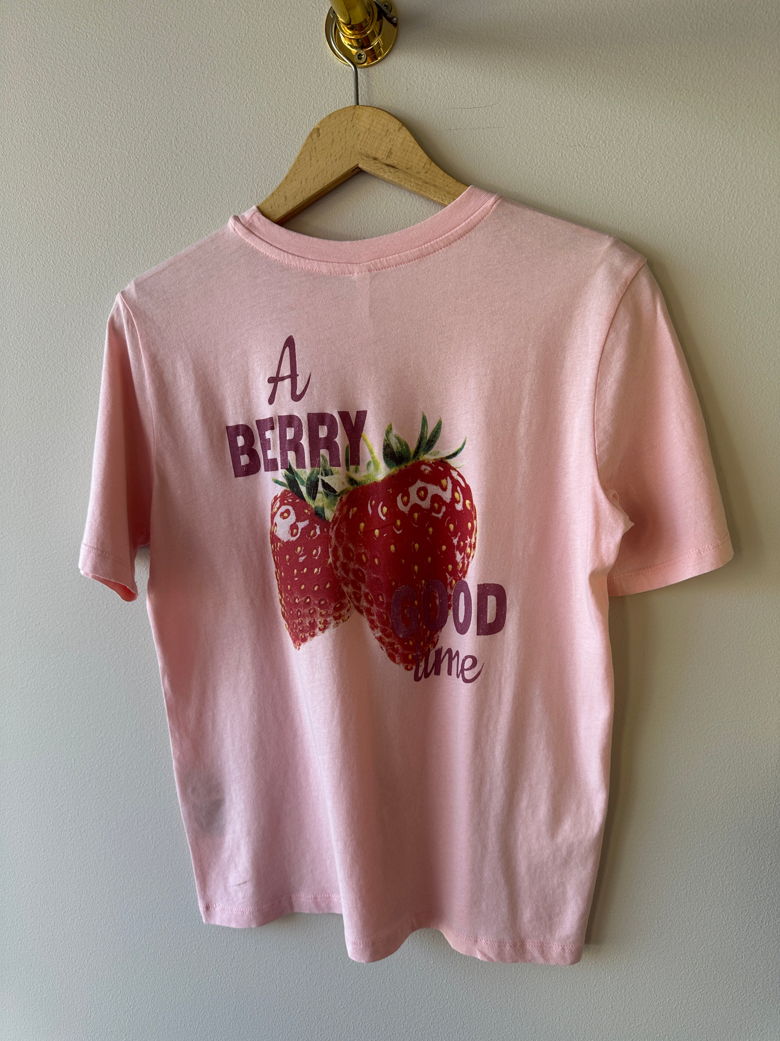 Baksida av rosa oversized t-shirt med stort tryck av två röda jordgubbar och texten A Berry Good Time i mörkrött, hängande på galge mot beige vägg.