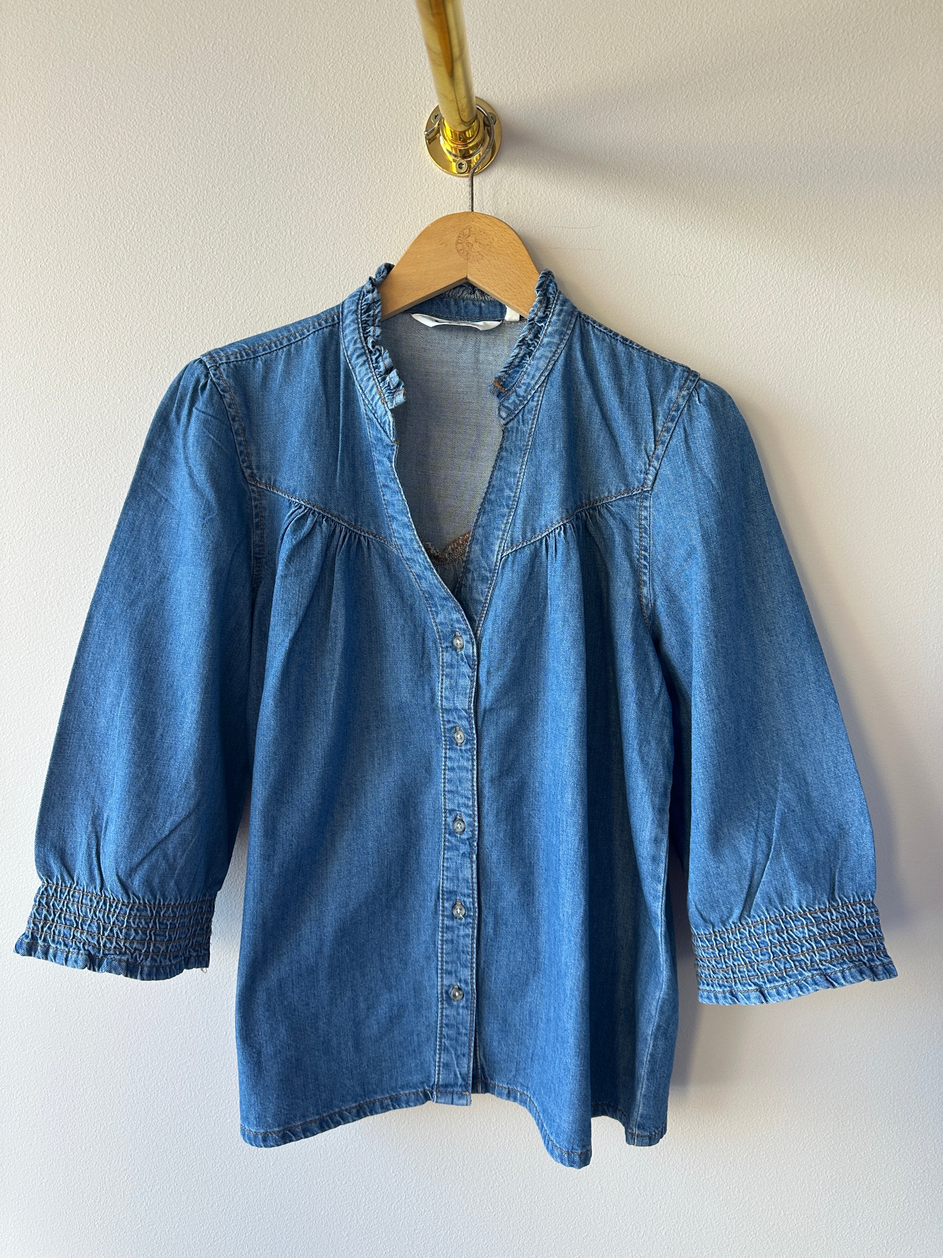 Mellanblå oversized denimskjorta med djup v-ringning, volangkrage och puffiga 3/4-ärmar med smockade manschetter hängande på galge mot vit vägg.