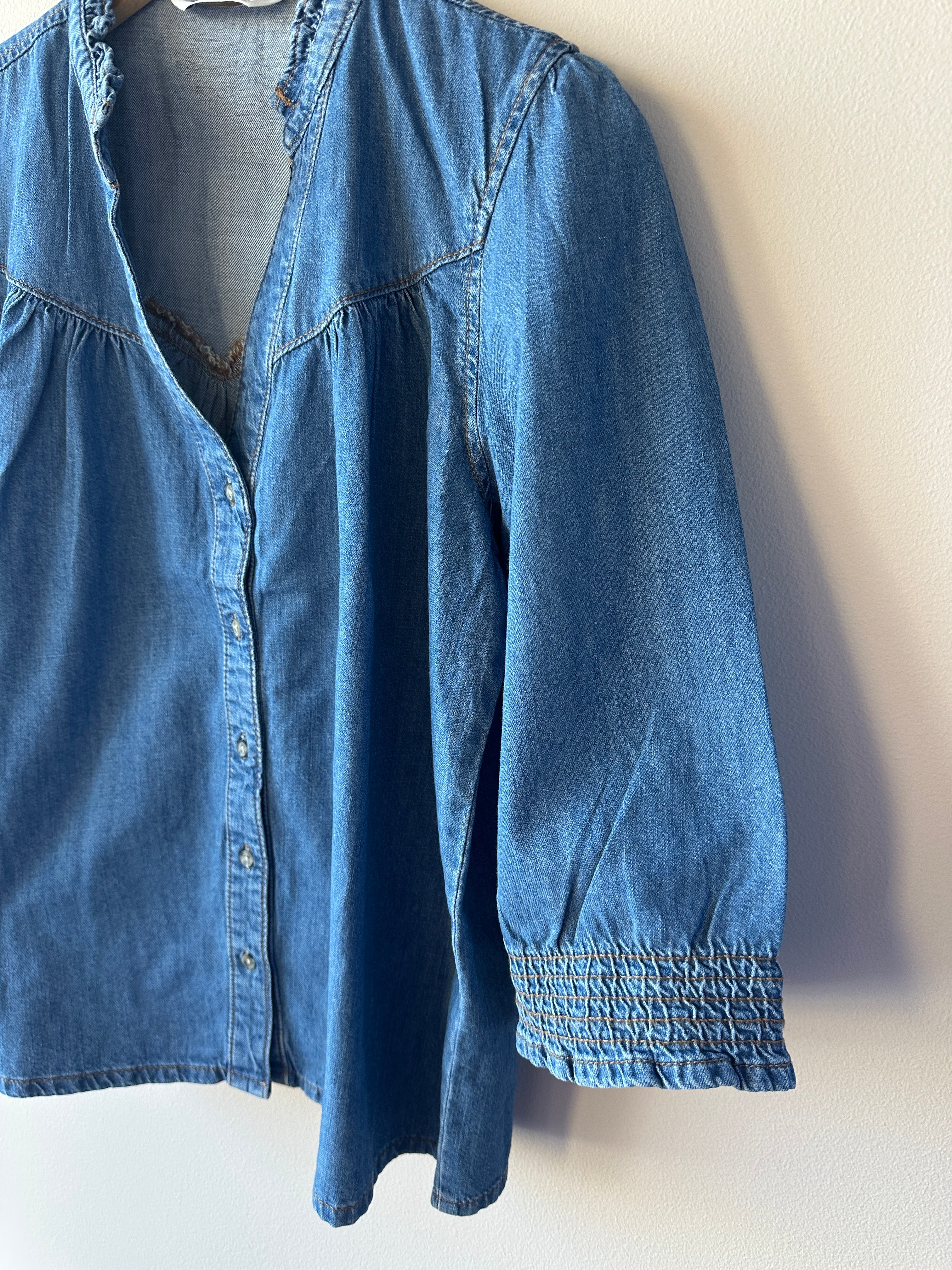 Närbild på mellanblå denimskjorta som visar volangkrage med v-ringning och puffig ärm med smockad manschett, hängande på galge mot vit vägg.