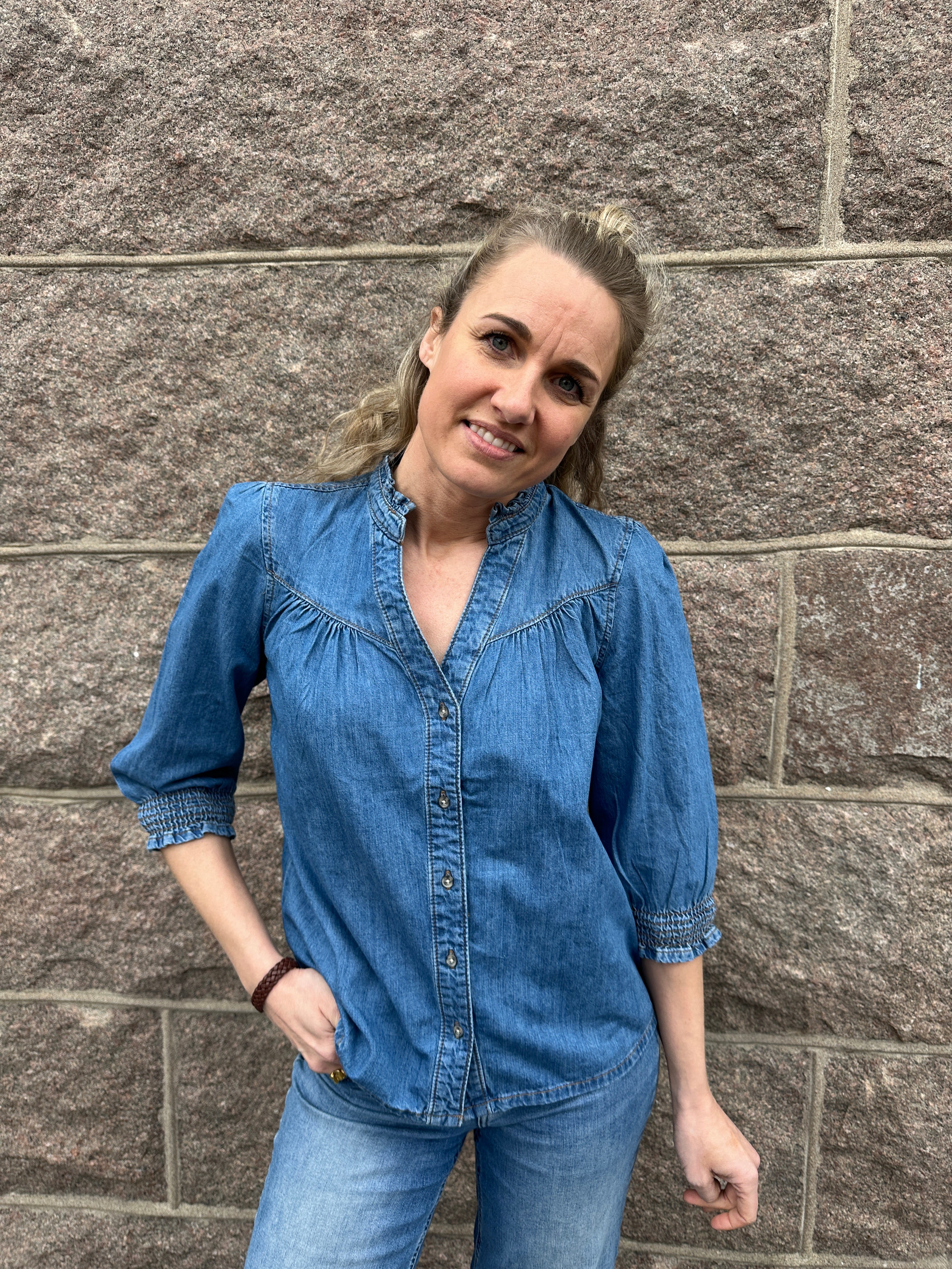 Kvinna iklädd mellanblå jeansskjorta med volangkrage, v-ringning, knappar fram och trekvartslånga puffärmar med smockade manschetter, poserar framför grå stenvägg.