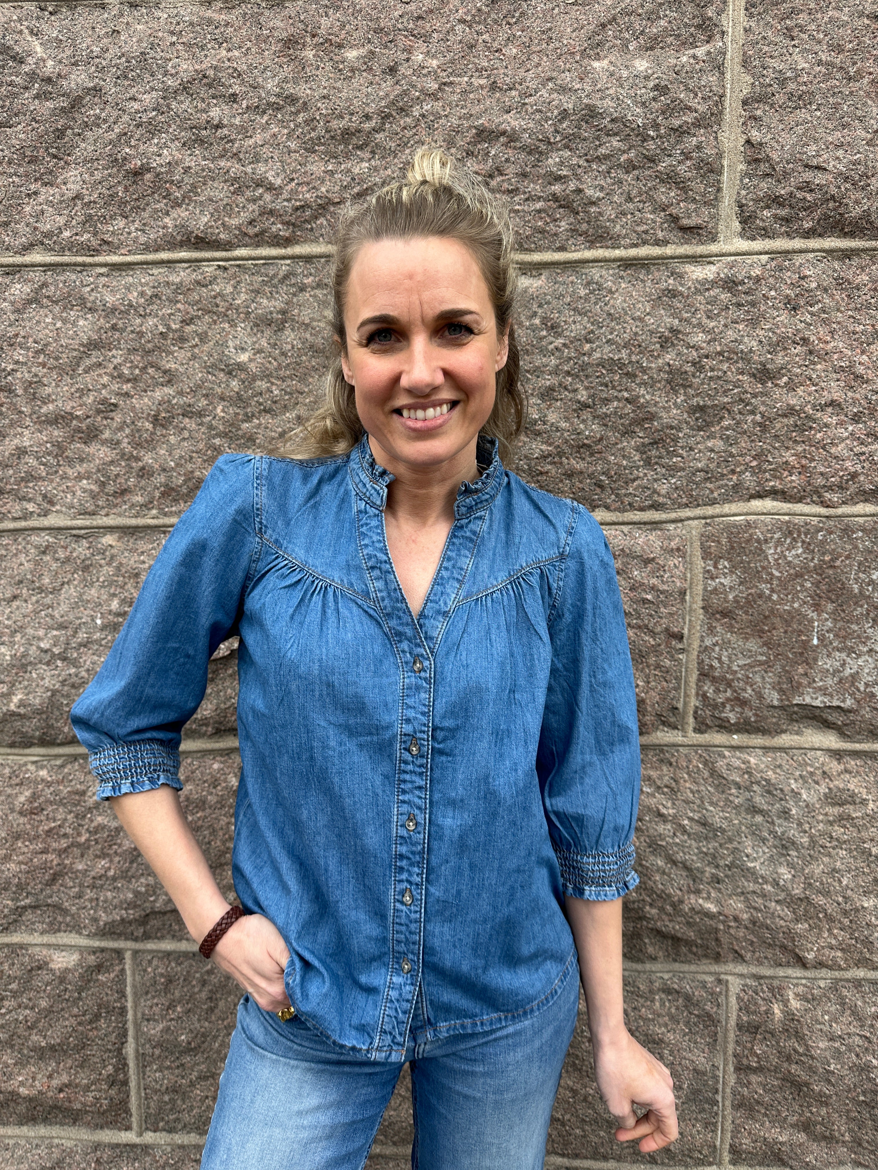 Kvinna iklädd mellanblå jeansskjorta med volangkrage, v-ringning och trekvartslånga puffärmar med smockade manschetter, handen i fickan stående framför grå stenvägg.