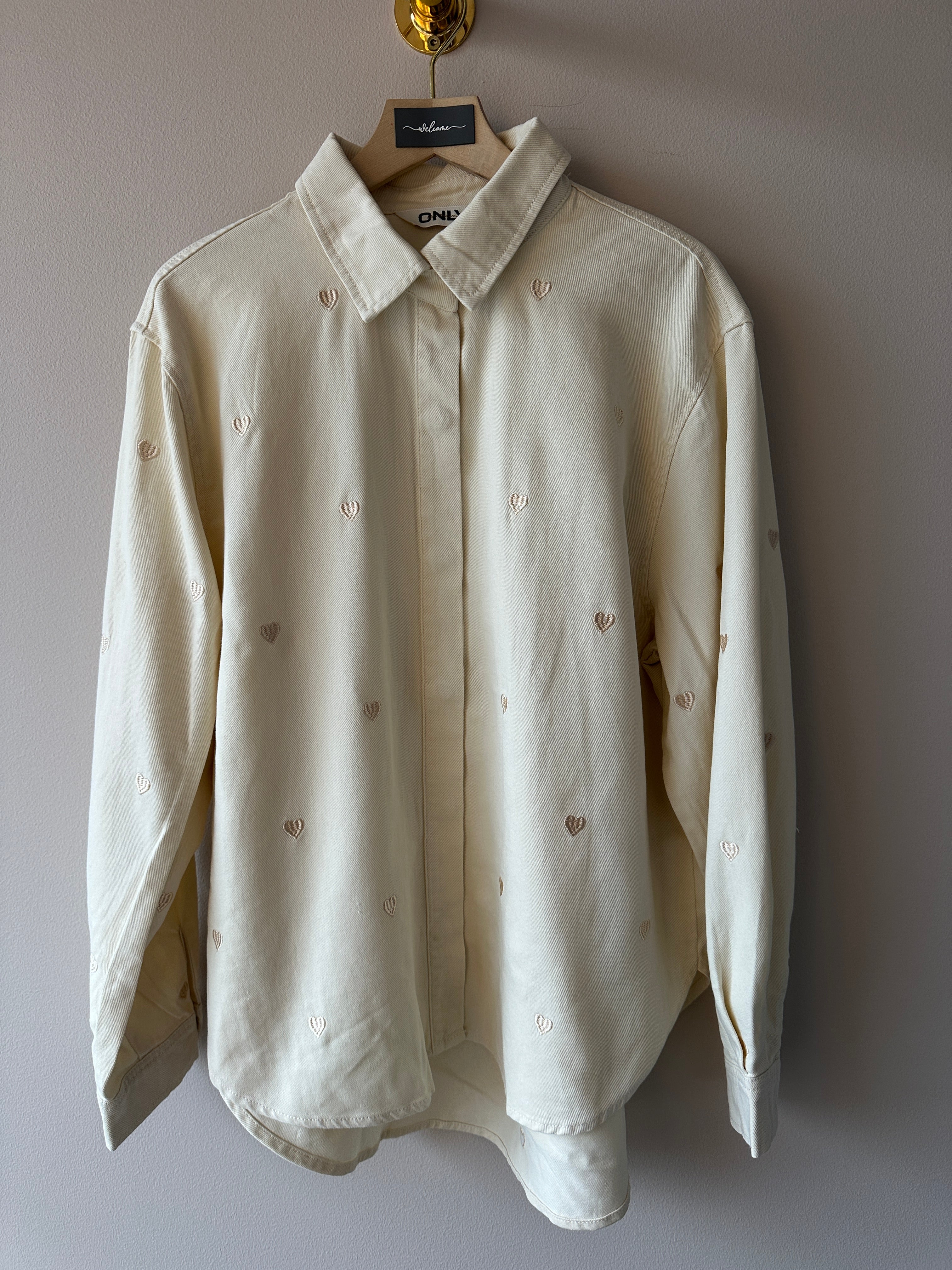 Crèmevit oversized skjorta med broderade hjärtan, hängd på galge mot ljusgrå vägg.