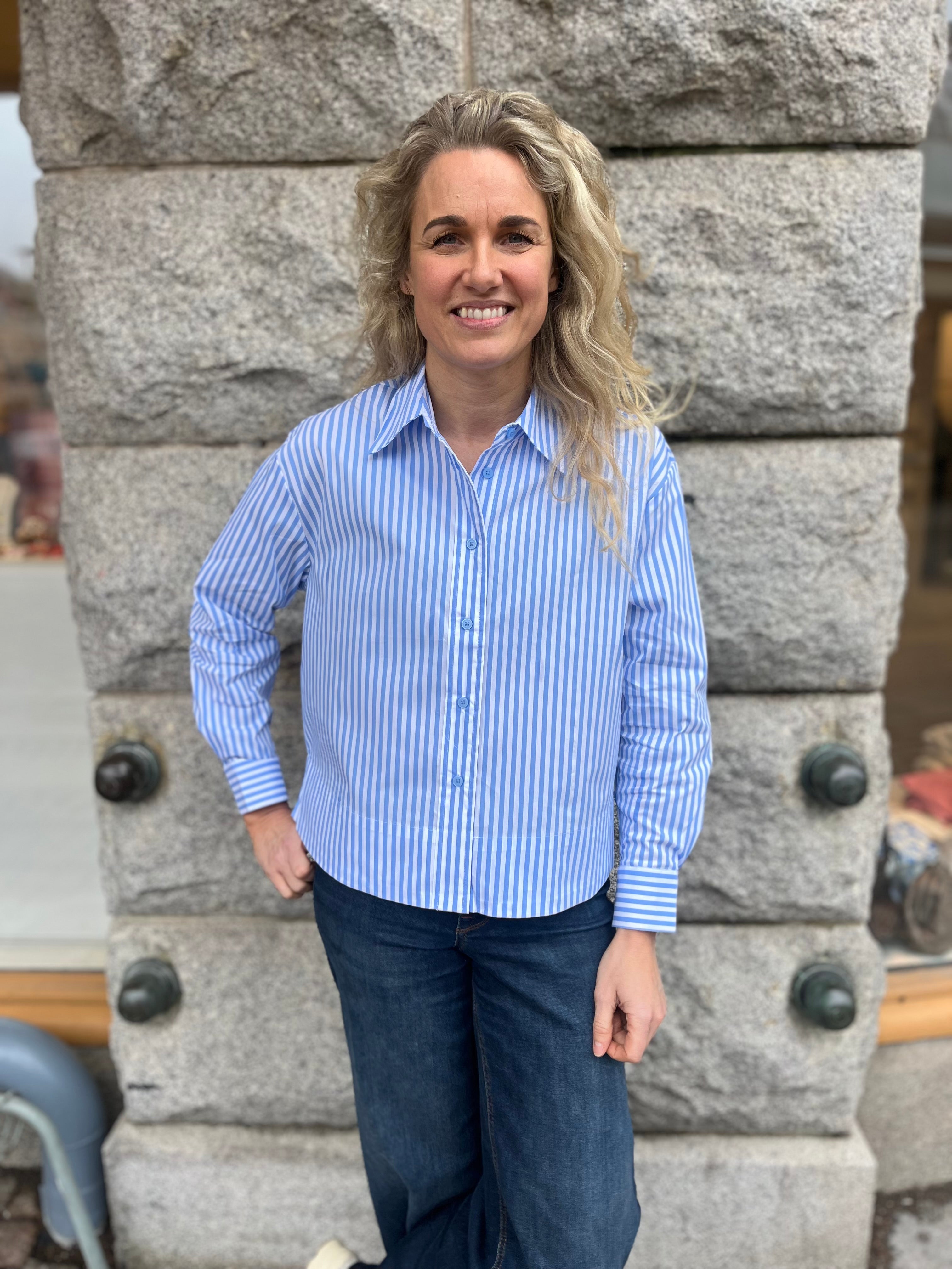 Blåvitrandig damskjorta med lång ärm och knappar, buret med mörkblå jeans – modell poserar framför stenvägg utomhus.