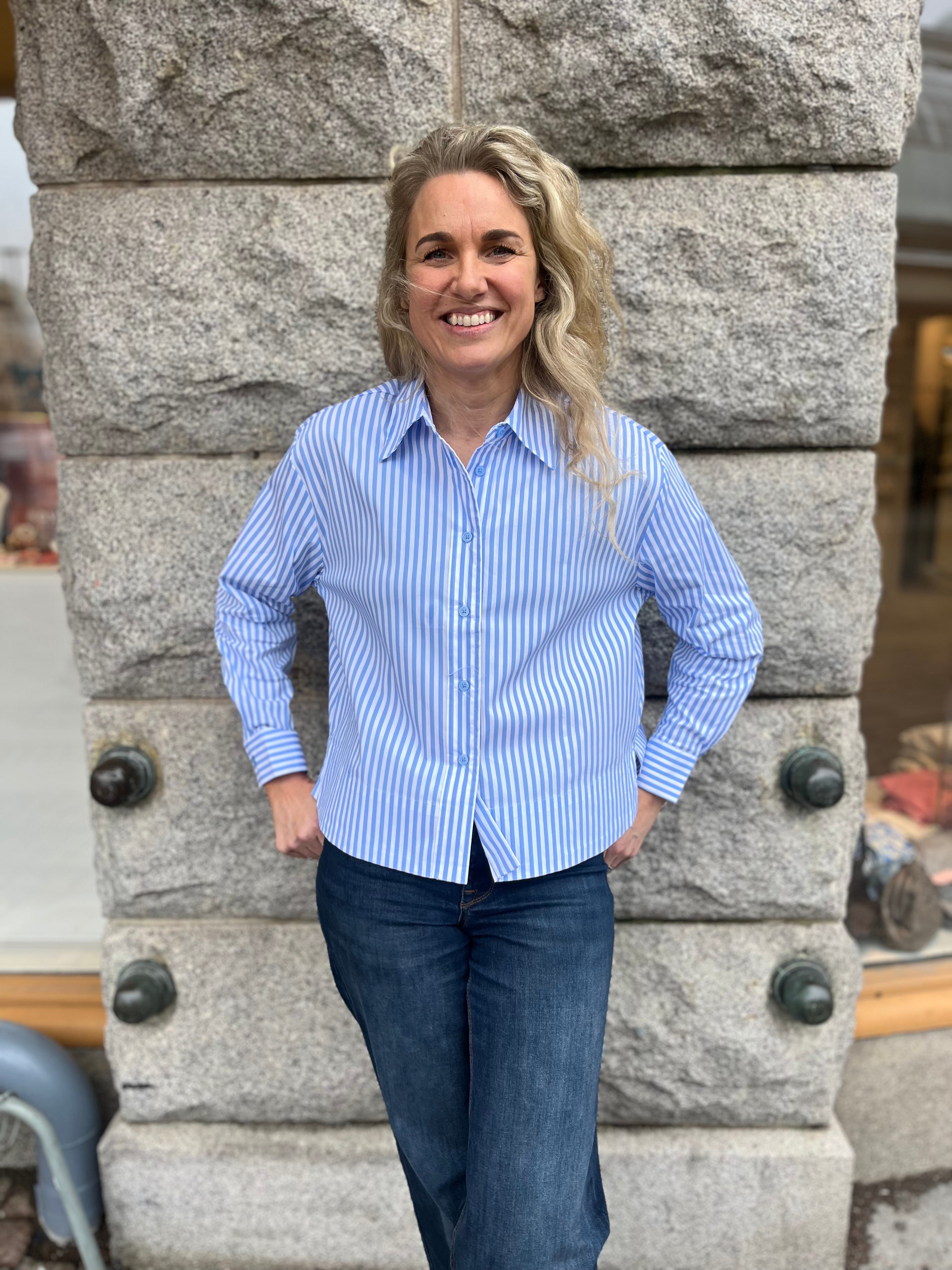 Kvinna i blåvitrandig skjorta med knappar och klassisk krage, stylade med mörkblå jeans – fotograferad utomhus mot en grå stenvägg.