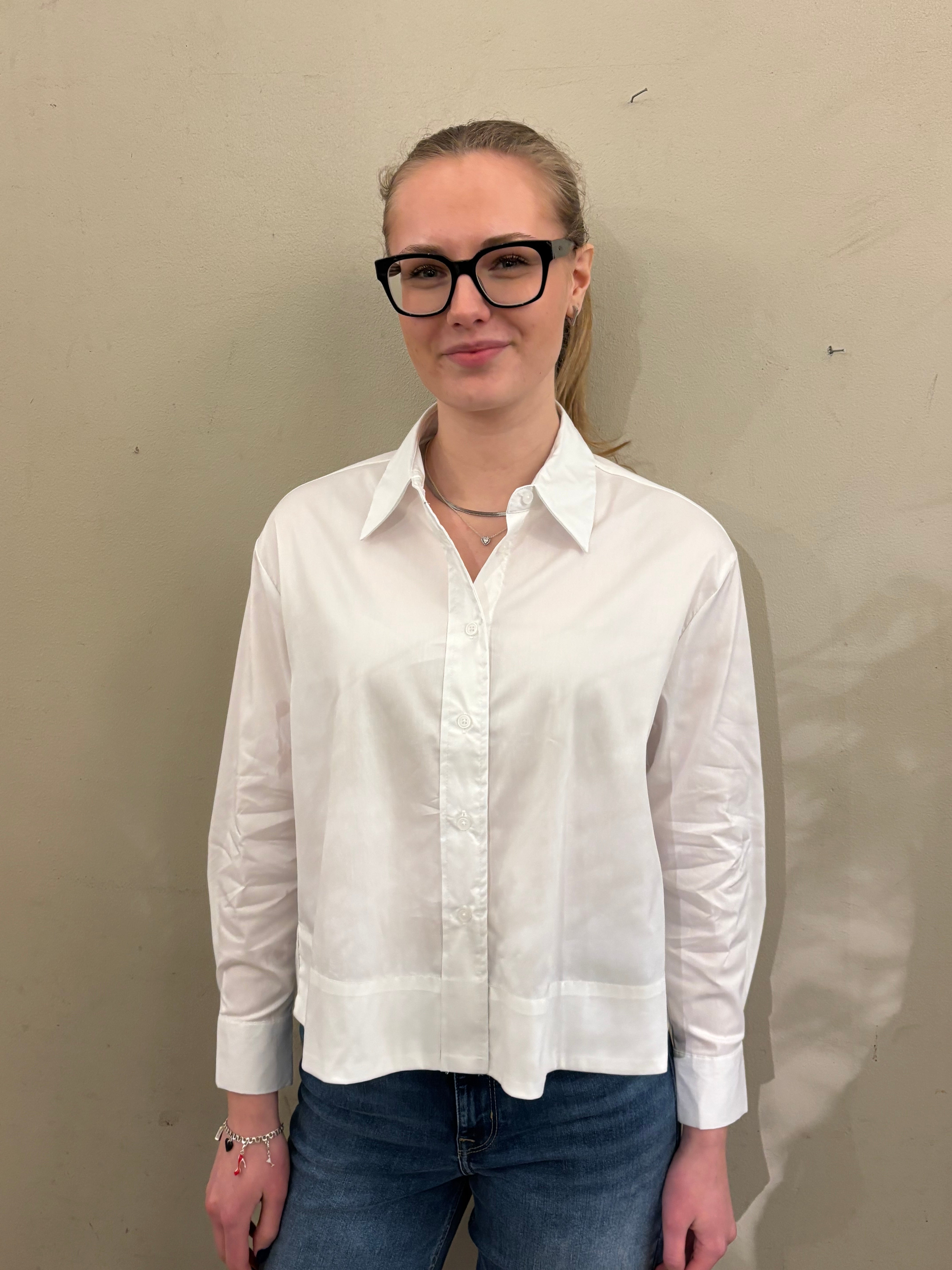 Helbild framifrån av kvinna i vit strukturerad skjorta med klassisk krage och breda manschetter, stylade med blå jeans mot beige vägg.