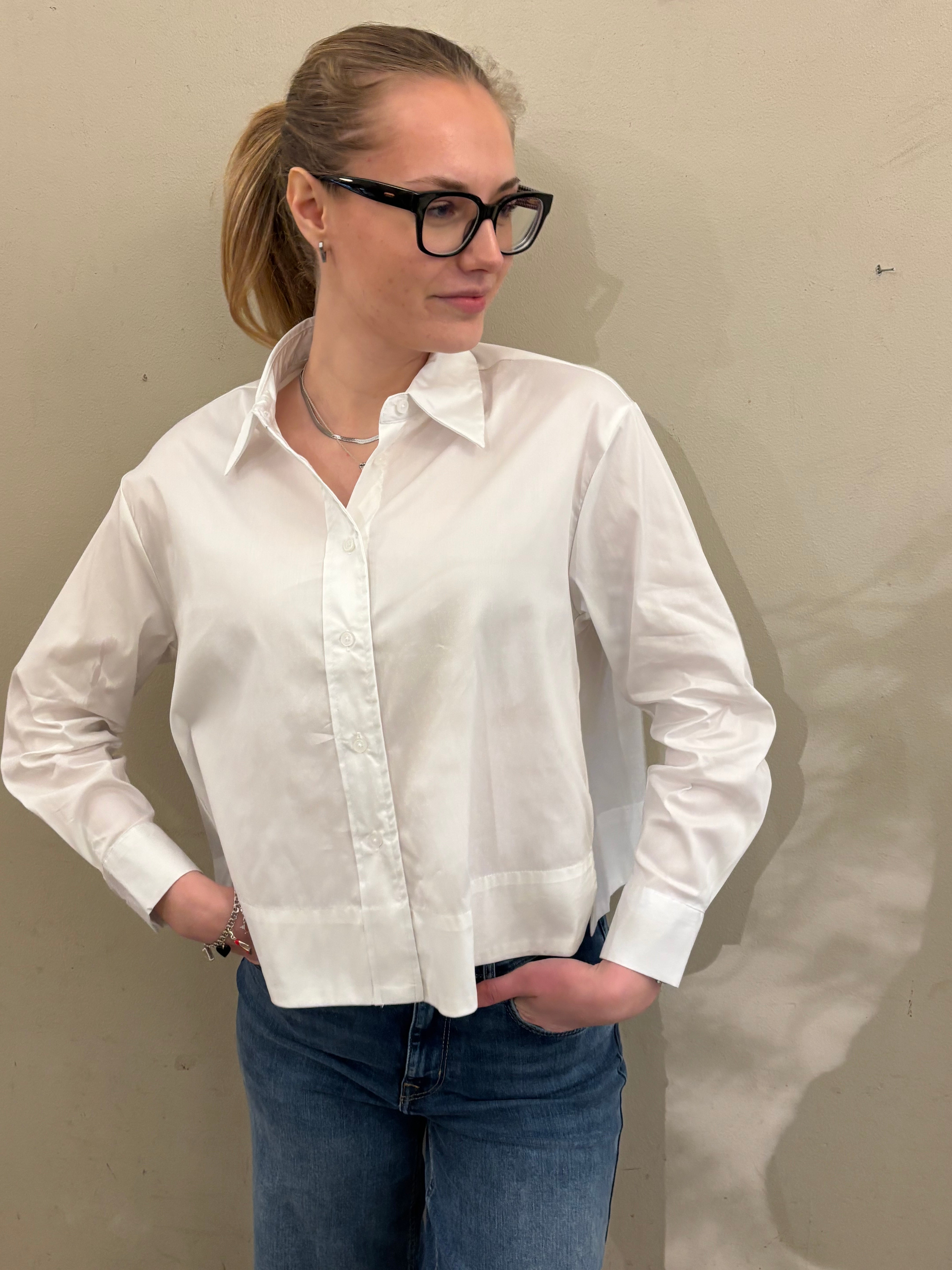 Kvinna i kortare vit skjorta med klassisk krage, dolda knappar och breda manschetter, stylade med blå jeans mot beige vägg.