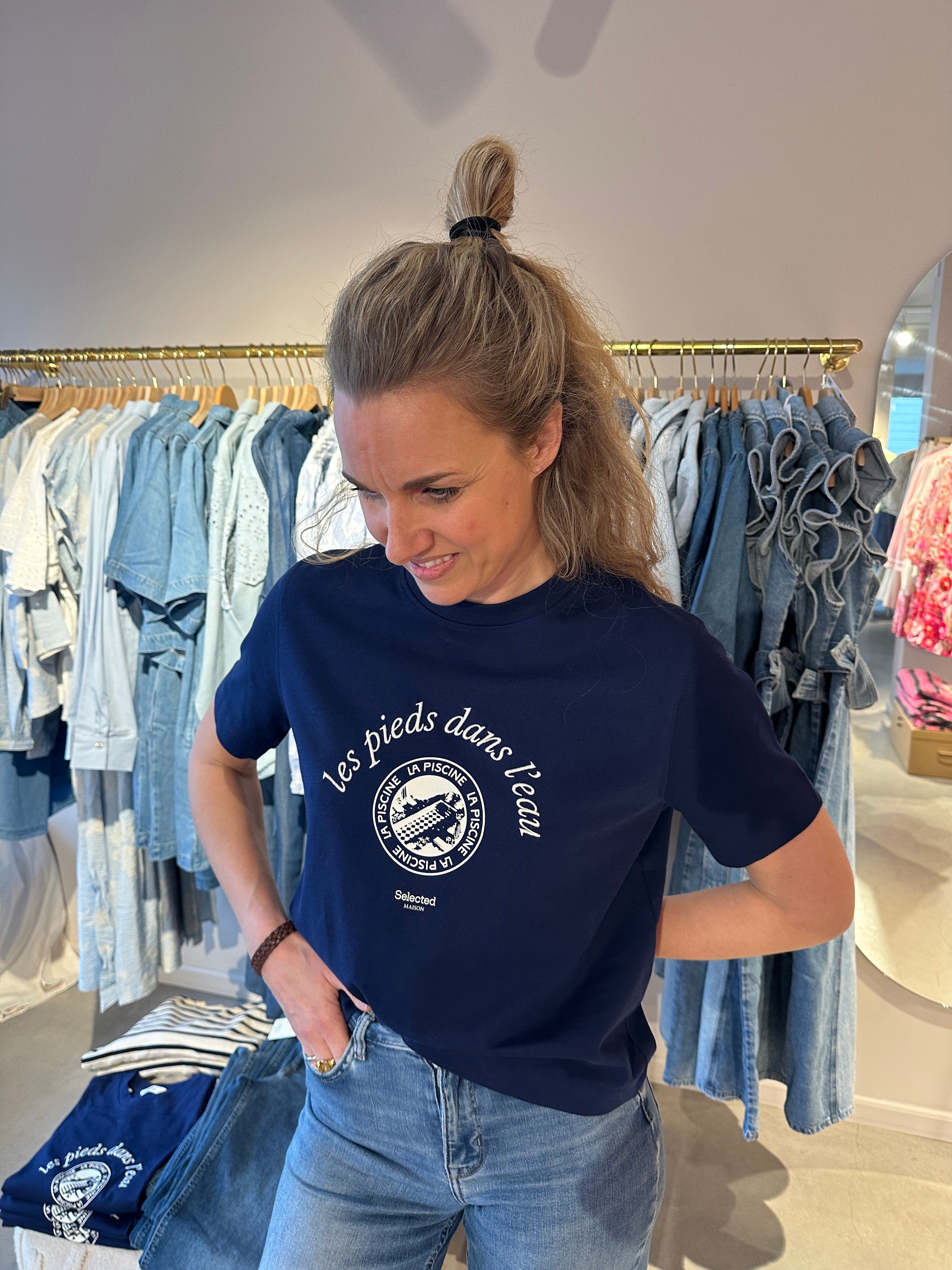 Kvinna iklädd marinblå t-shirt med vitt grafiskt tryck – texten les pieds dans l'eau och runt La Piscine-emblem med poolmotiv på bröstet, bärs med blå jeans i butiksmiljö i bakgrunden.