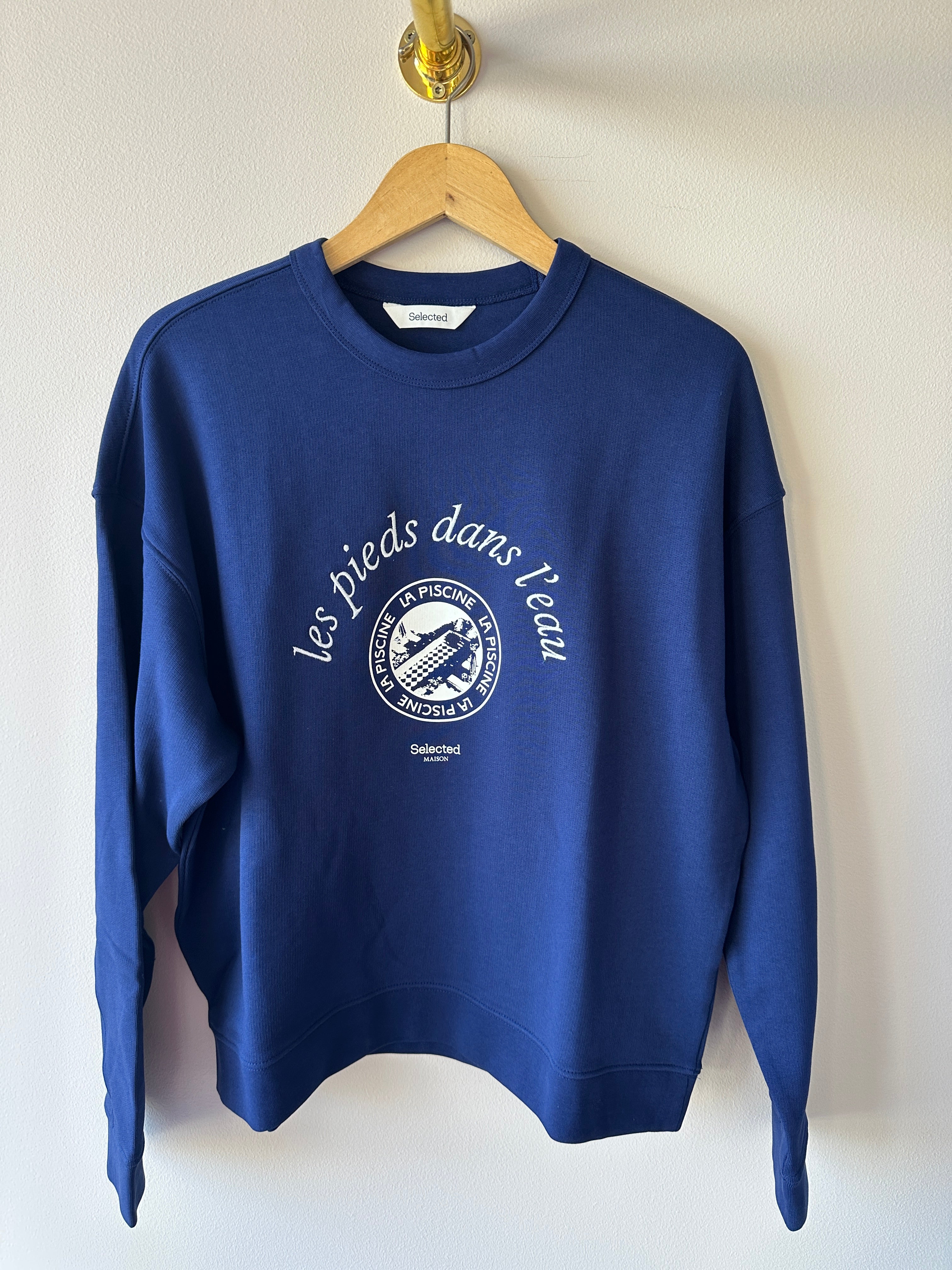Marinblå oversized sweatshirt med vitt tryck av texten les pieds dans l'eau och La Piscine-motiv på bröstet, rund hals och långa ärmar hängande på galge mot vit vägg.