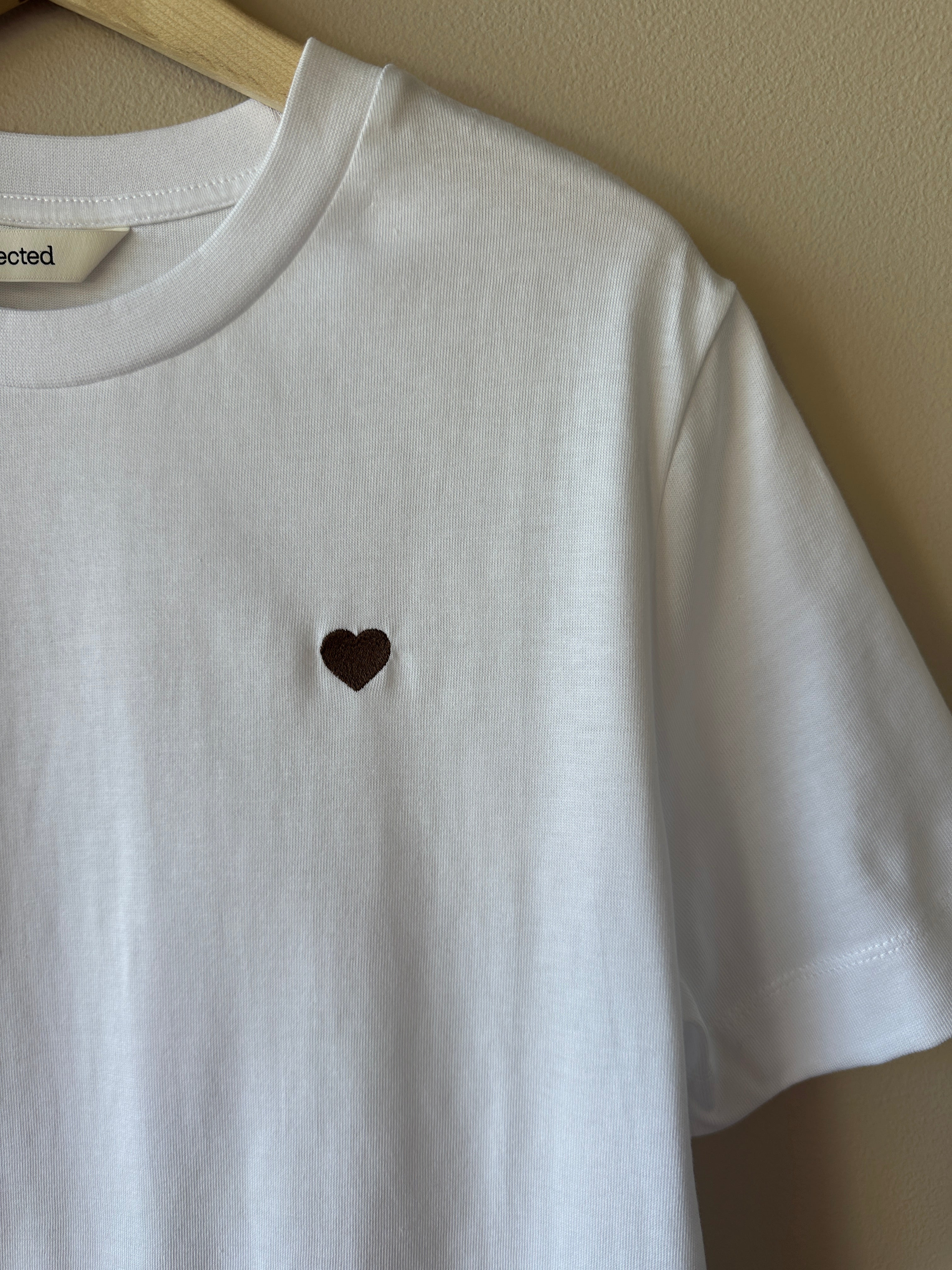 Närbild på bröstet av vit t-shirt som visar litet brunt hjärtbroderi på mjukt vitt bomullstyg, etikett med texten Selected synlig i halsen.