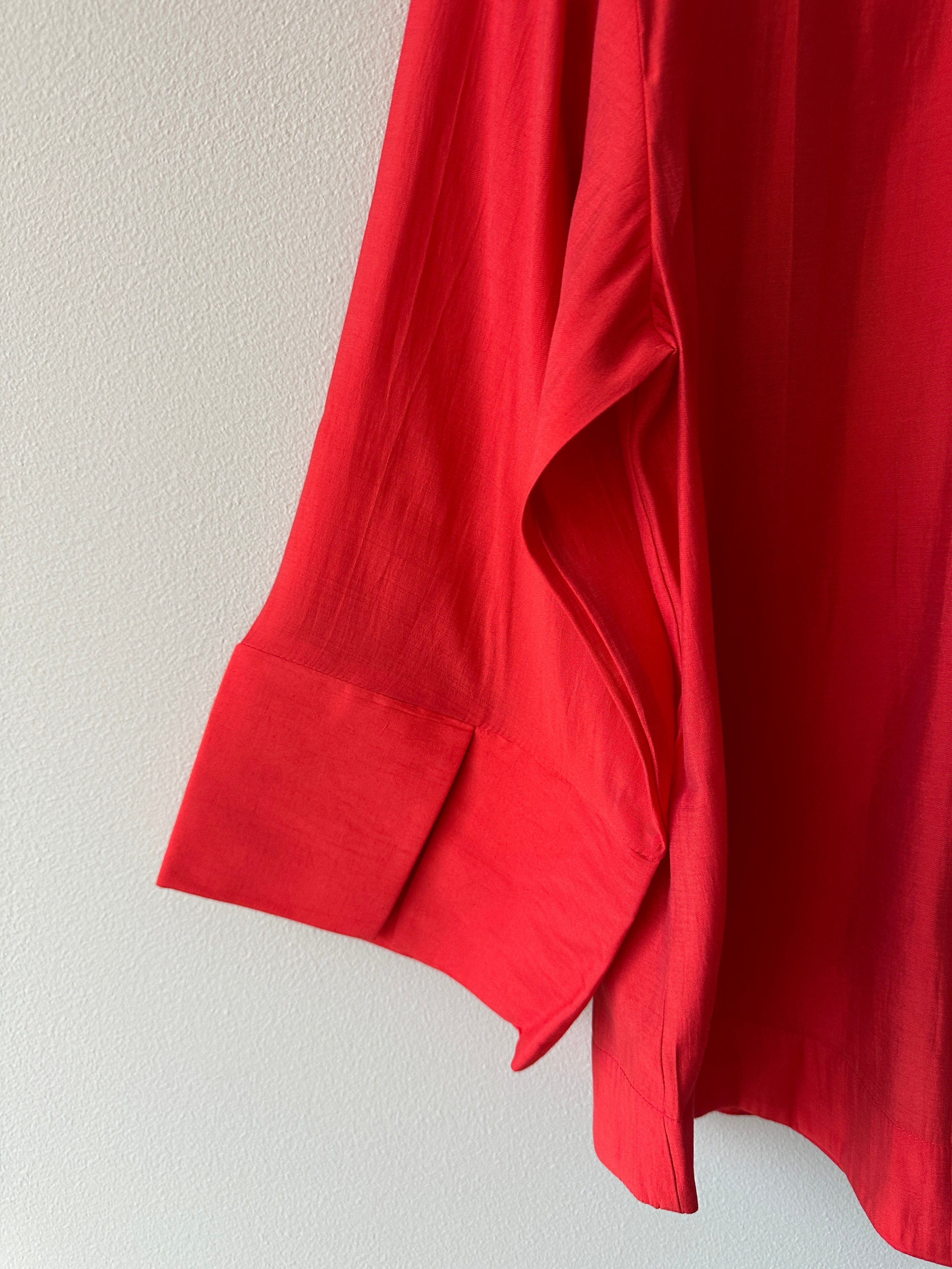 Närbild på ärmslut och sidosläpp på klarröd skjorta i lätt flytande tyg med bred ärmkant.