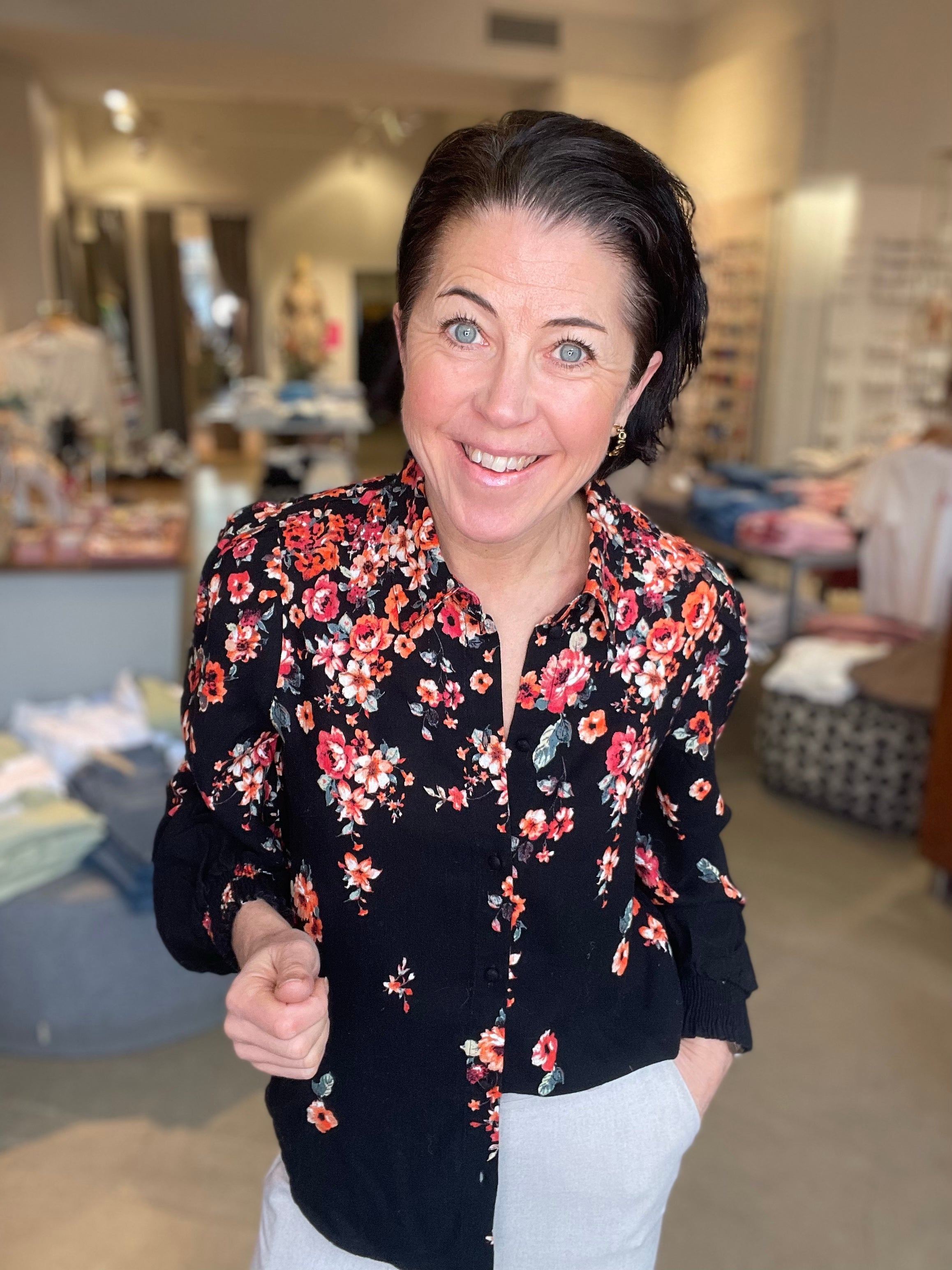 Kvinna iklädd svart skjorta med rött och rosa blommigt tryck och långa ärmar med ribbade muddar, ler brett mot kameran i butiksmiljö.
