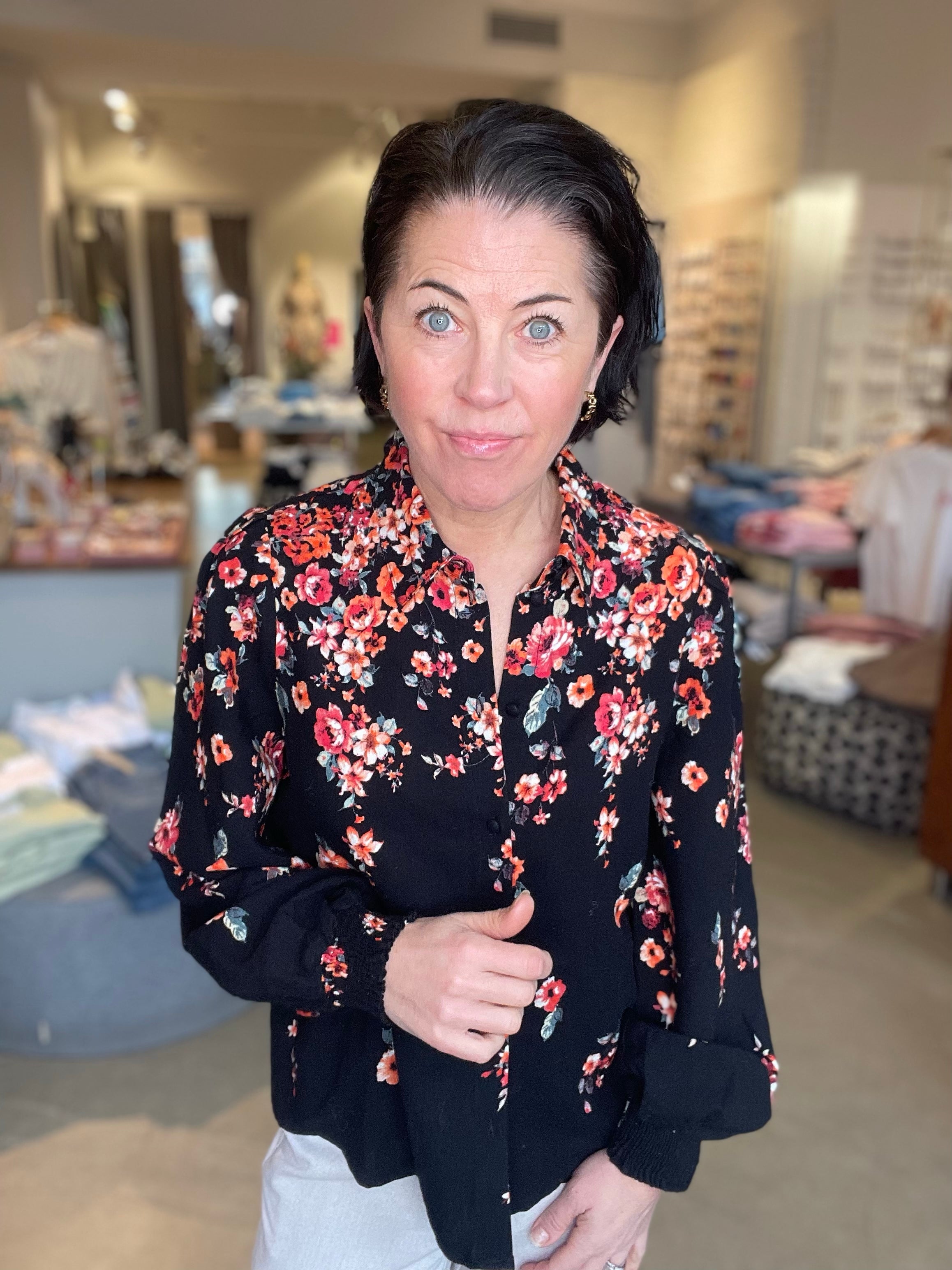 Kvinna iklädd svart skjorta med rött och rosa blommigt tryck, v-ringad krage med knappar fram och långa ärmar, håller i kragen och tittar in i kameran i butiksmiljö.