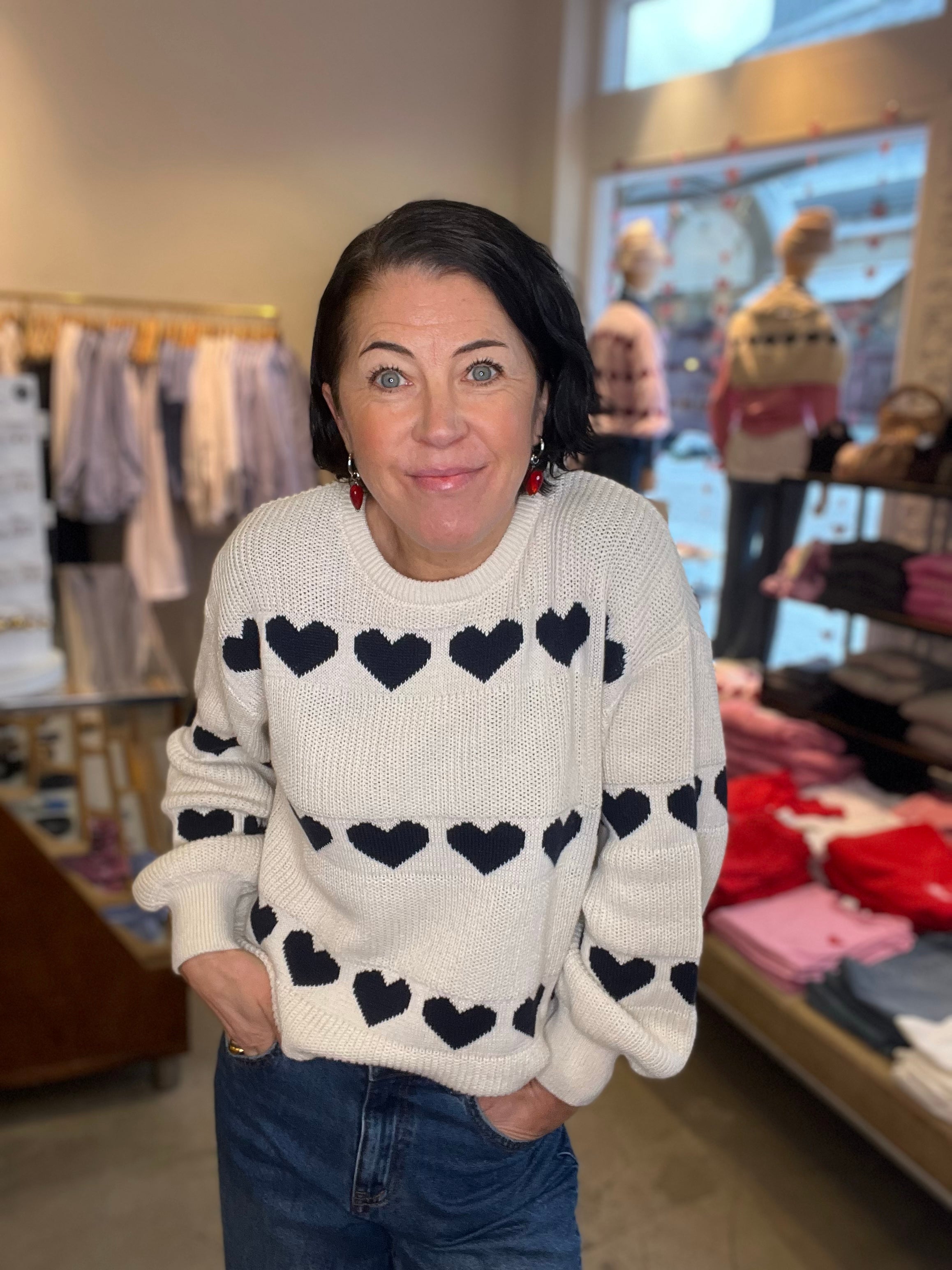 Kvinna iklädd vit jacquardstickad tröja med stora marinblå hjärtan i rader över hela tyget, rund hals och långa ärmar, poserar med händerna i fickorna i butiksmiljö.