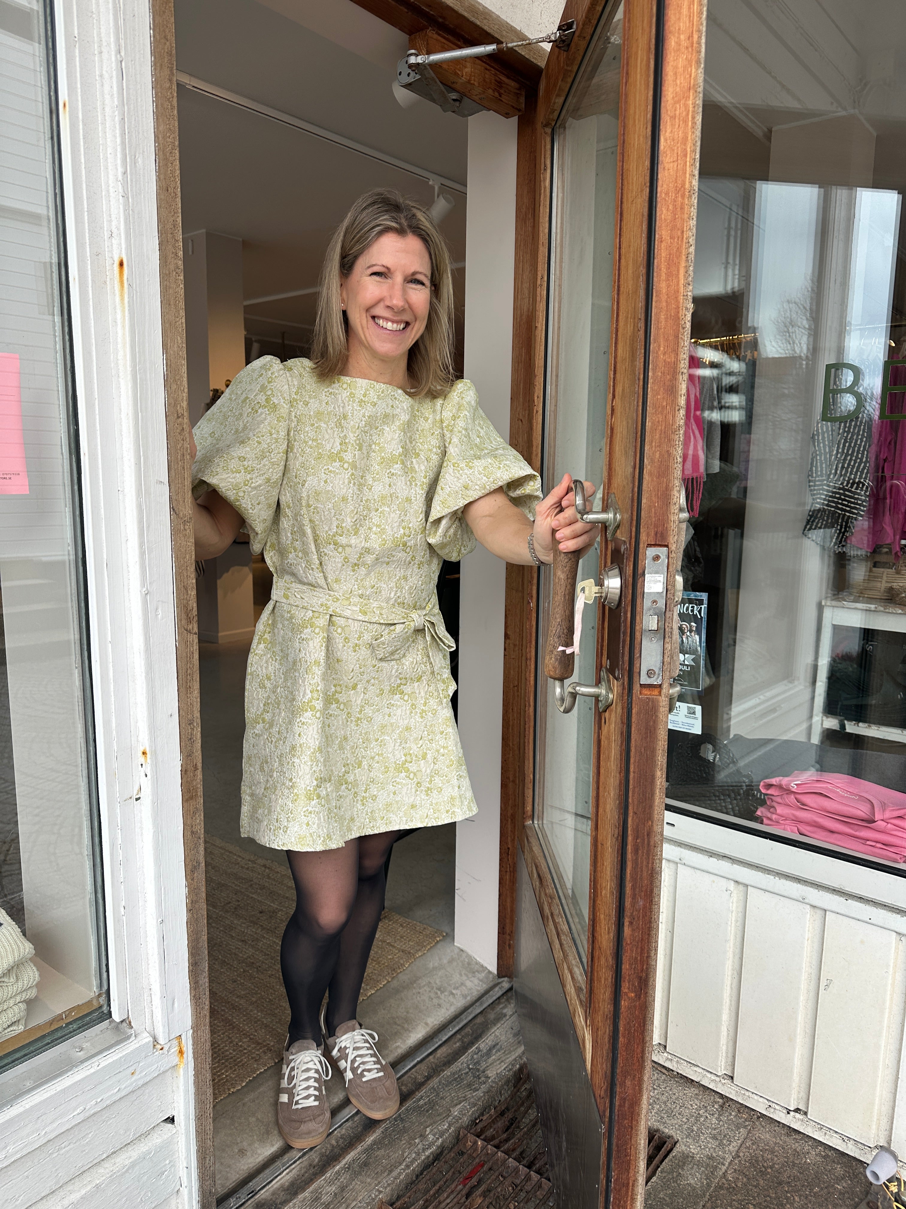 Leende kvinna i helbild i kort ljusgrön jacquardklänning med båtringning, volangärmar och knytskärp i midjan, stylade med grå strumpbyxor och sneakers poserar i butiksdörren.,