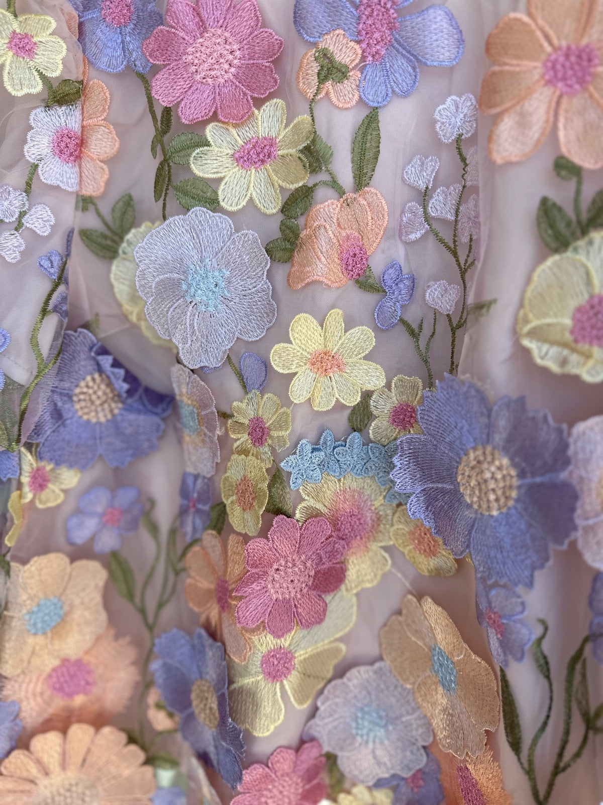 Närbild på tyget av lavendelfärgad topp som visar detaljerade 3D-broderade pastellblommor i lila, rosa, gult, orange och blått i varierande storlekar på genomskinligt mesh.