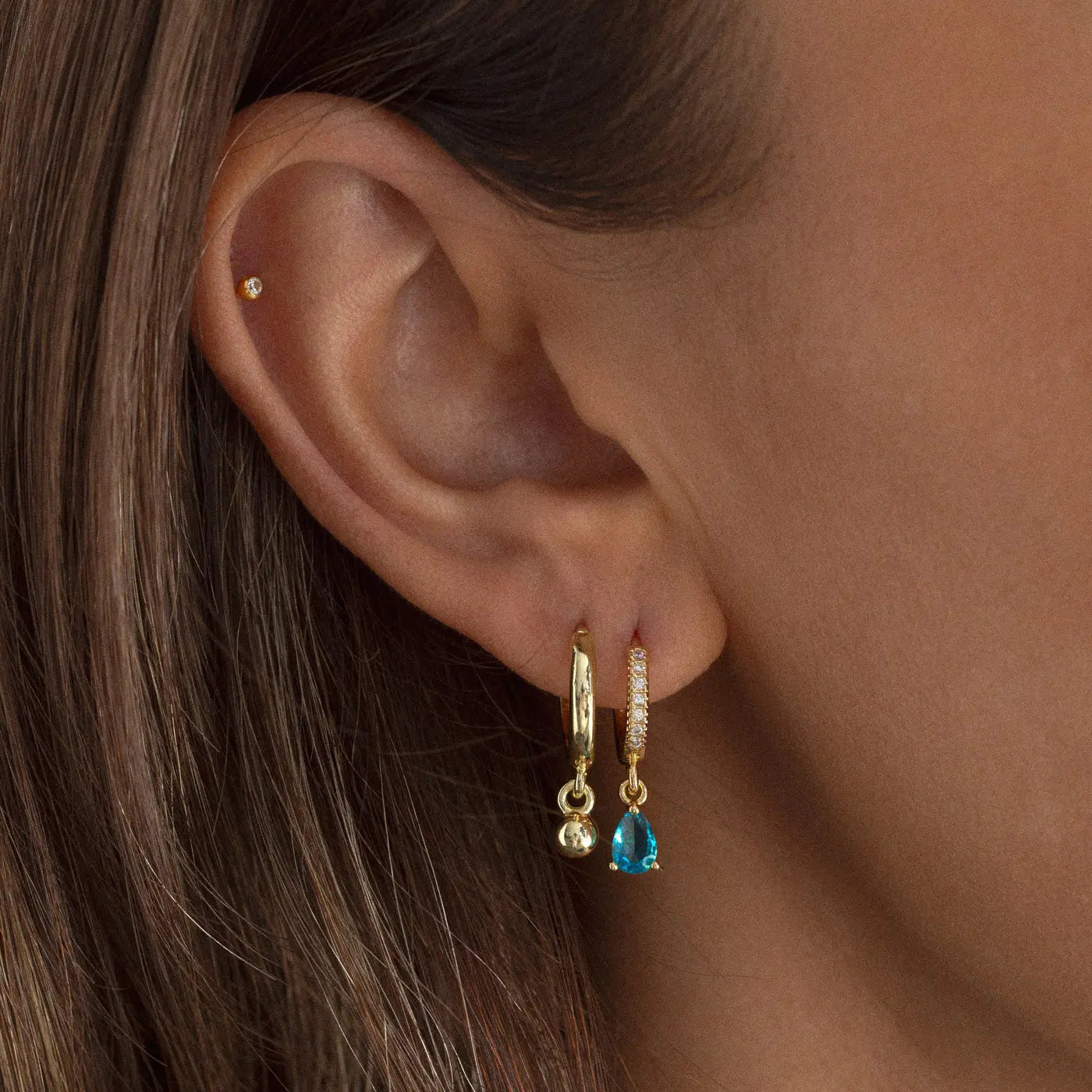 Kailo - Ball Hoop Earrings - BECCASIN STORE - Gold ball hoop örhänge with a small hanging blue gemstone smycke on a woman’s ear.