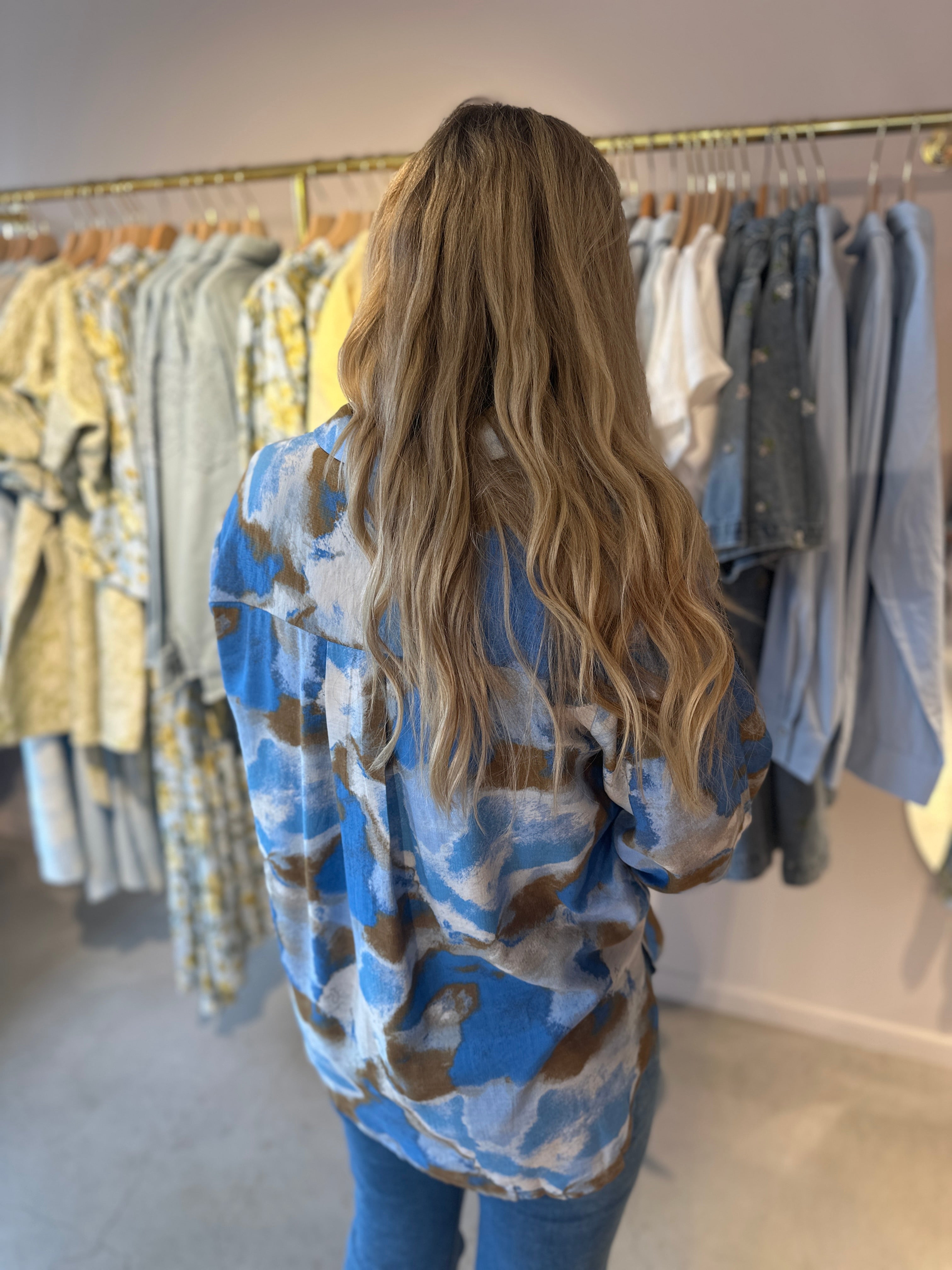 Kvinna bakifrån i oversized skjorta med abstrakt mönster i blå, khaki och vitt, stylade med blå jeans i butiksmiljö.